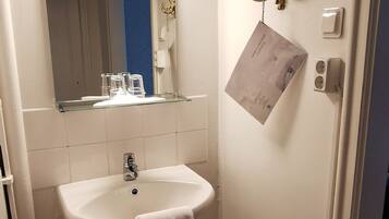 A small bathroom with a white sink, a mirror, a towel hanging on the wall, a small trash bin, and a light switch.