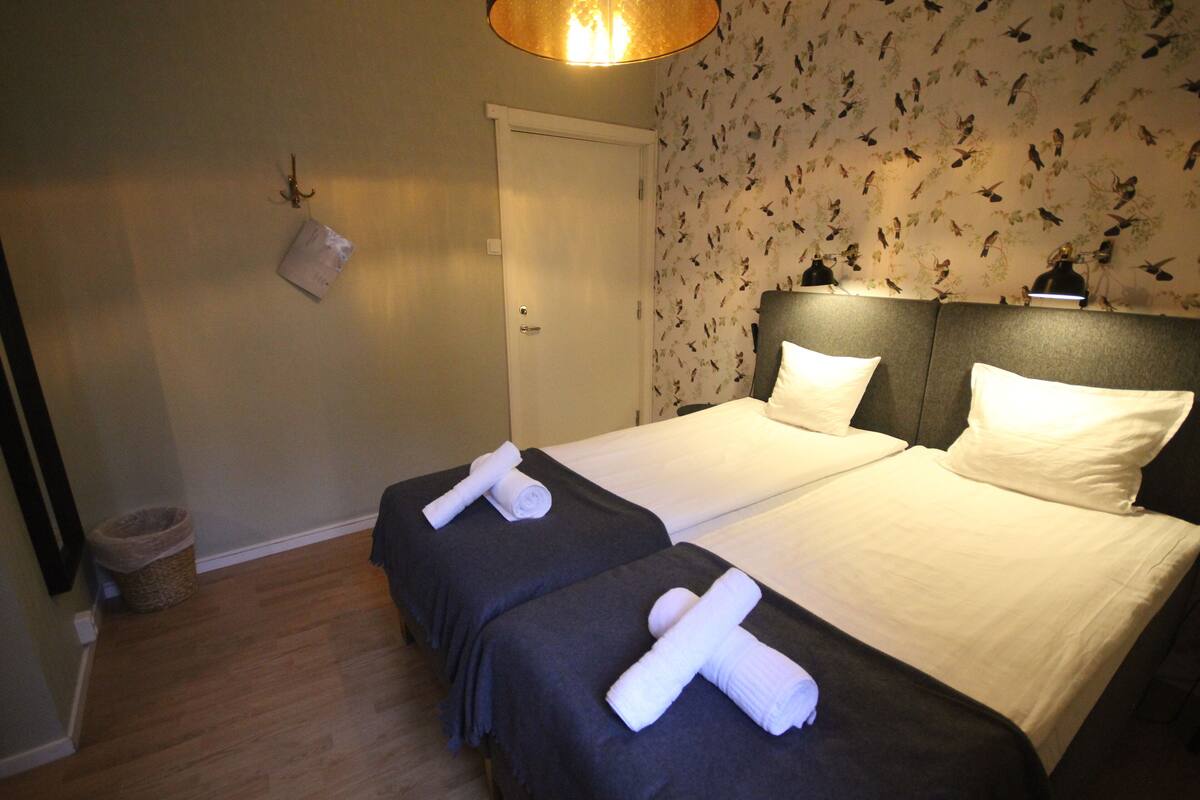 A hotel room with two beds, each with a dark headboard and white linens. There is a door with a light switch and a wall-mounted light fixture above it.