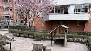 Un espacio al aire libre con arena, bancos de madera y una pequeña construcción de madera, rodeado por una cerca de metal y un edificio con amplias ventanas al fondo.