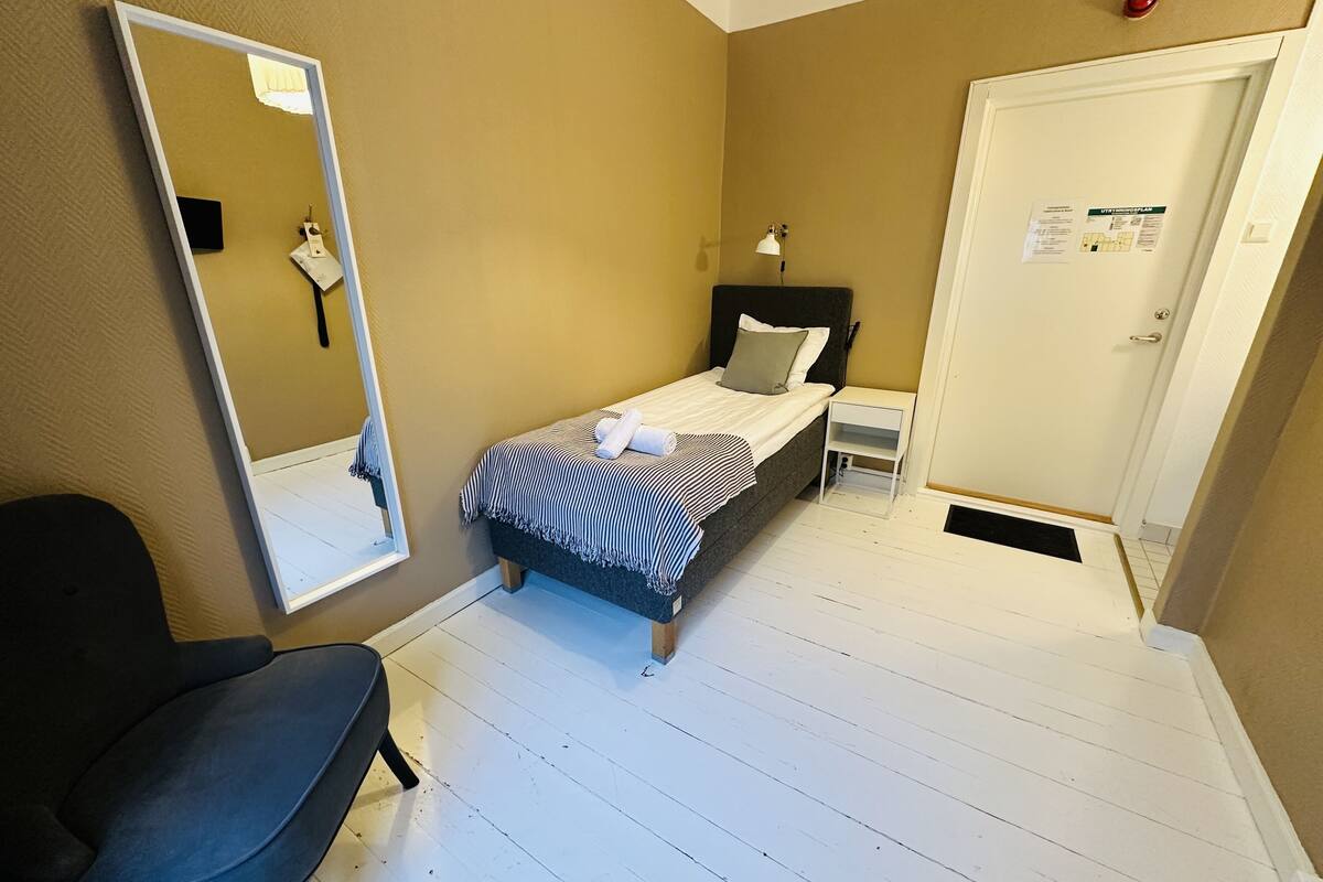 A bedroom with a bed, a chair, a bedside table, a nightstand, a mirror, and a door with a sign.