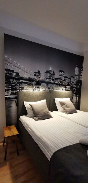 A bedroom with a large bed, two pillows, a nightstand, and a cityscape wall mural.