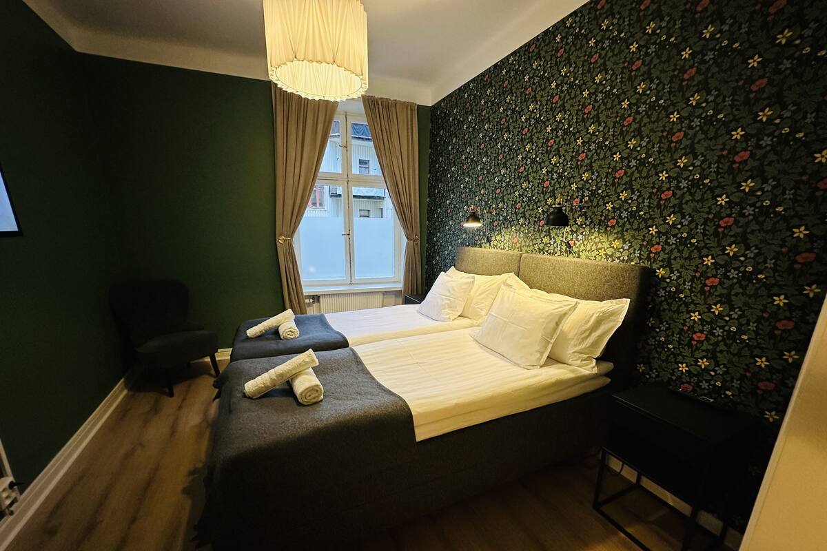 A double bed with white linens, a headboard with a floral pattern, and a window with curtains.