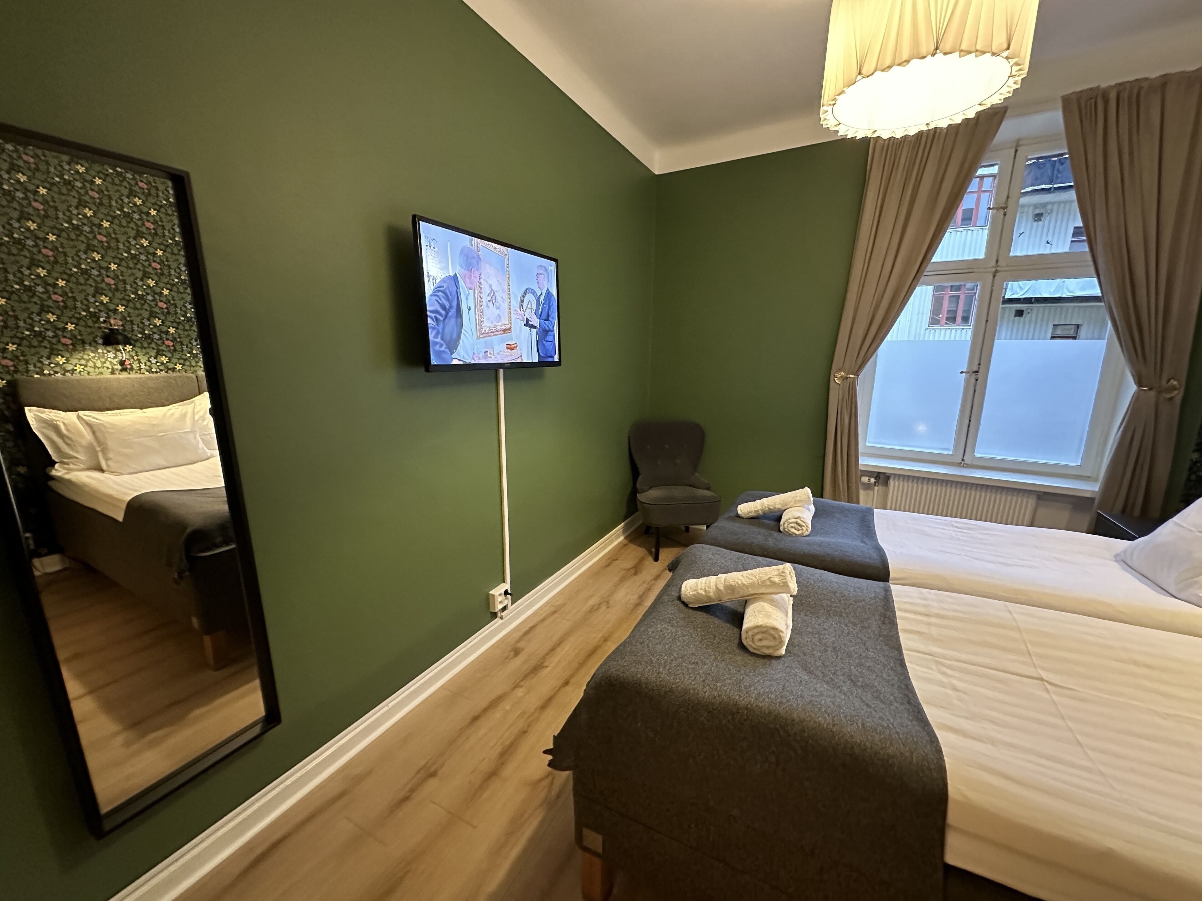Quarto de hotel com duas camas, televisão fixada na parede, uma cadeira e janela com cortinas.
