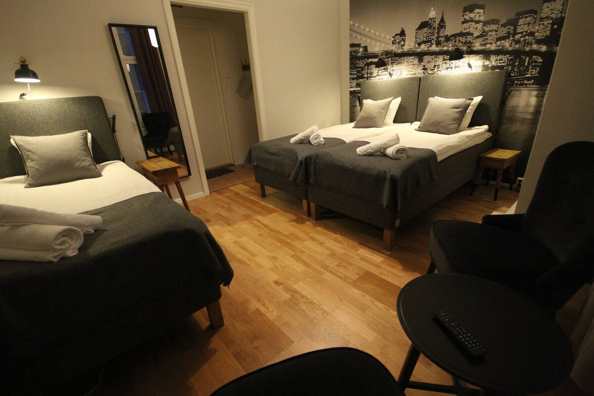 A hotel room with two beds, a chair, a small table, and a cityscape wall mural.