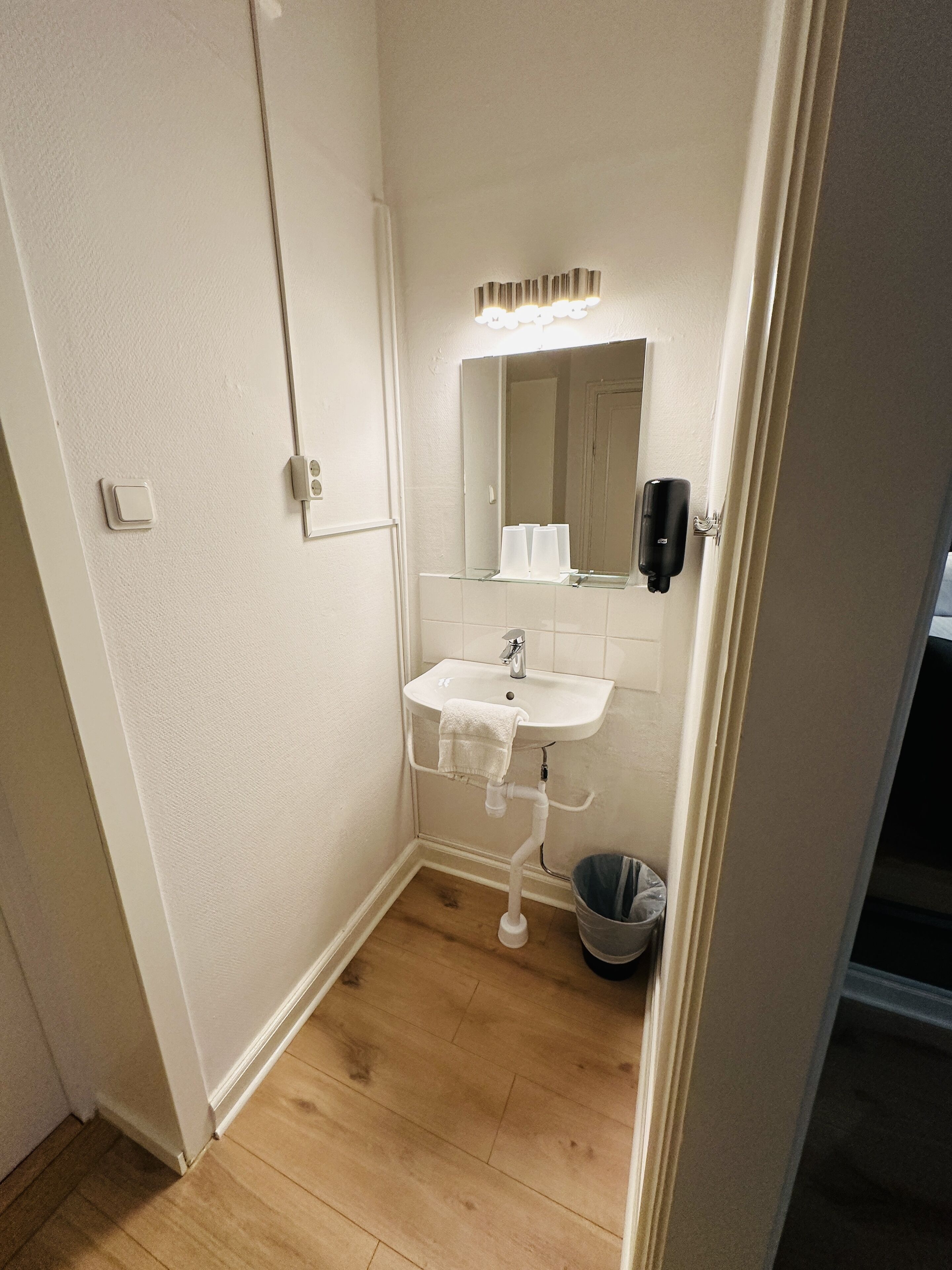 Un baño pequeño y bien iluminado con un lavamanos blanco, un espejo y un perchero para toallas.