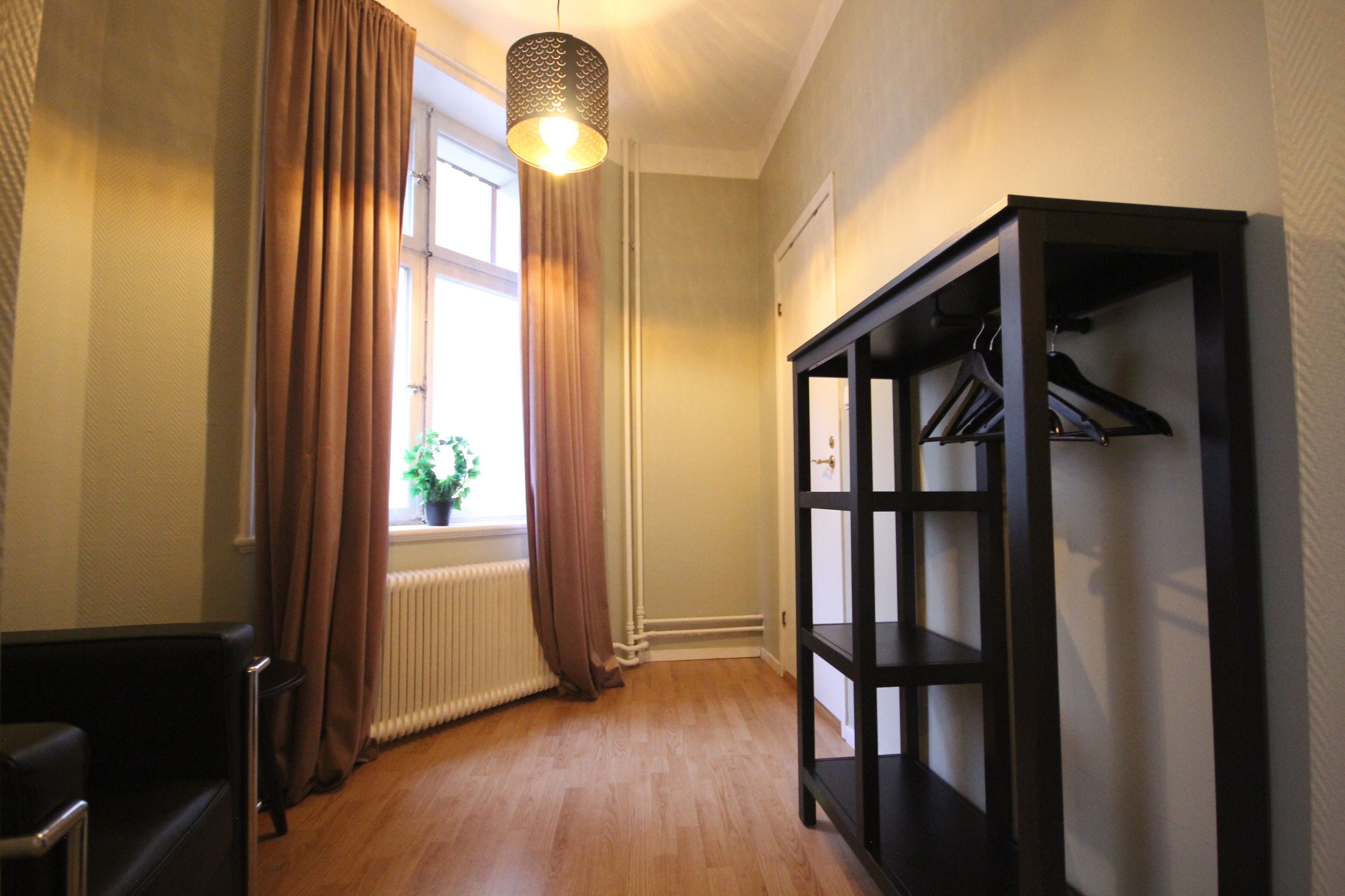 A hallway with a radiator, a hanging light fixture, a coat rack, and a window with curtains.