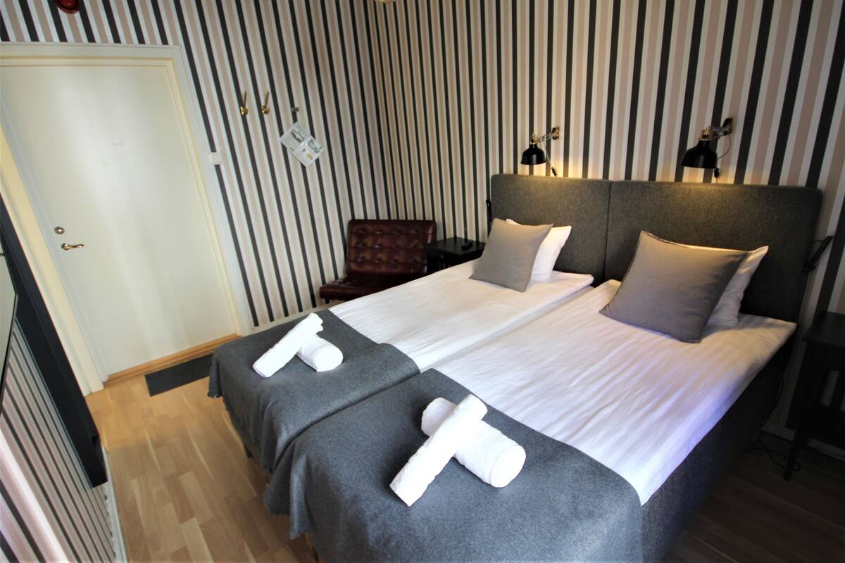 A hotel room with two beds, striped wallpaper, a wooden floor, a chair, and a nightstand with a lamp.