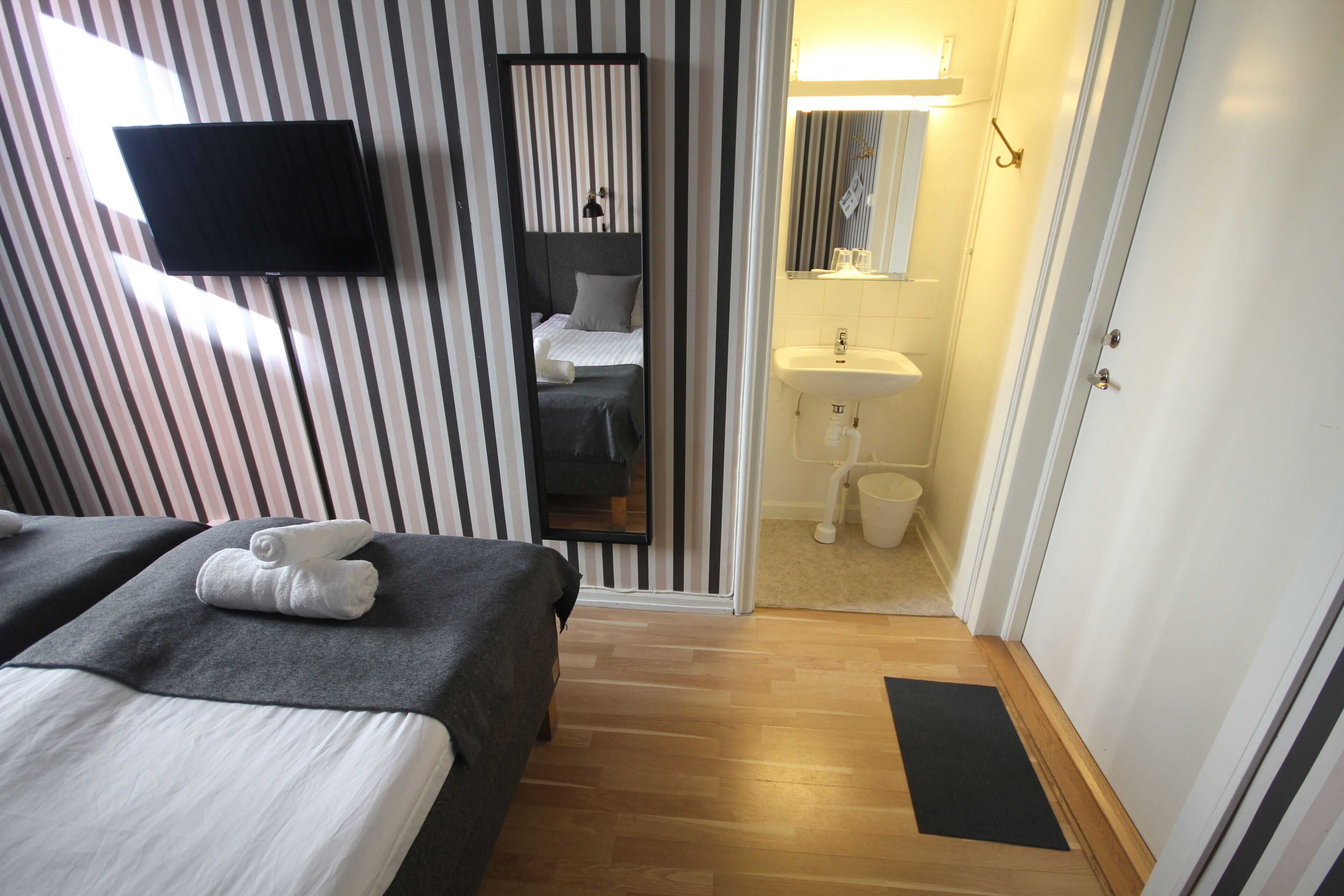 Habitación de hotel con papel tapiz a rayas, un televisor de pantalla plana, una cama con edredón blanco y gris, una mesita de noche pequeña y suelo de madera.