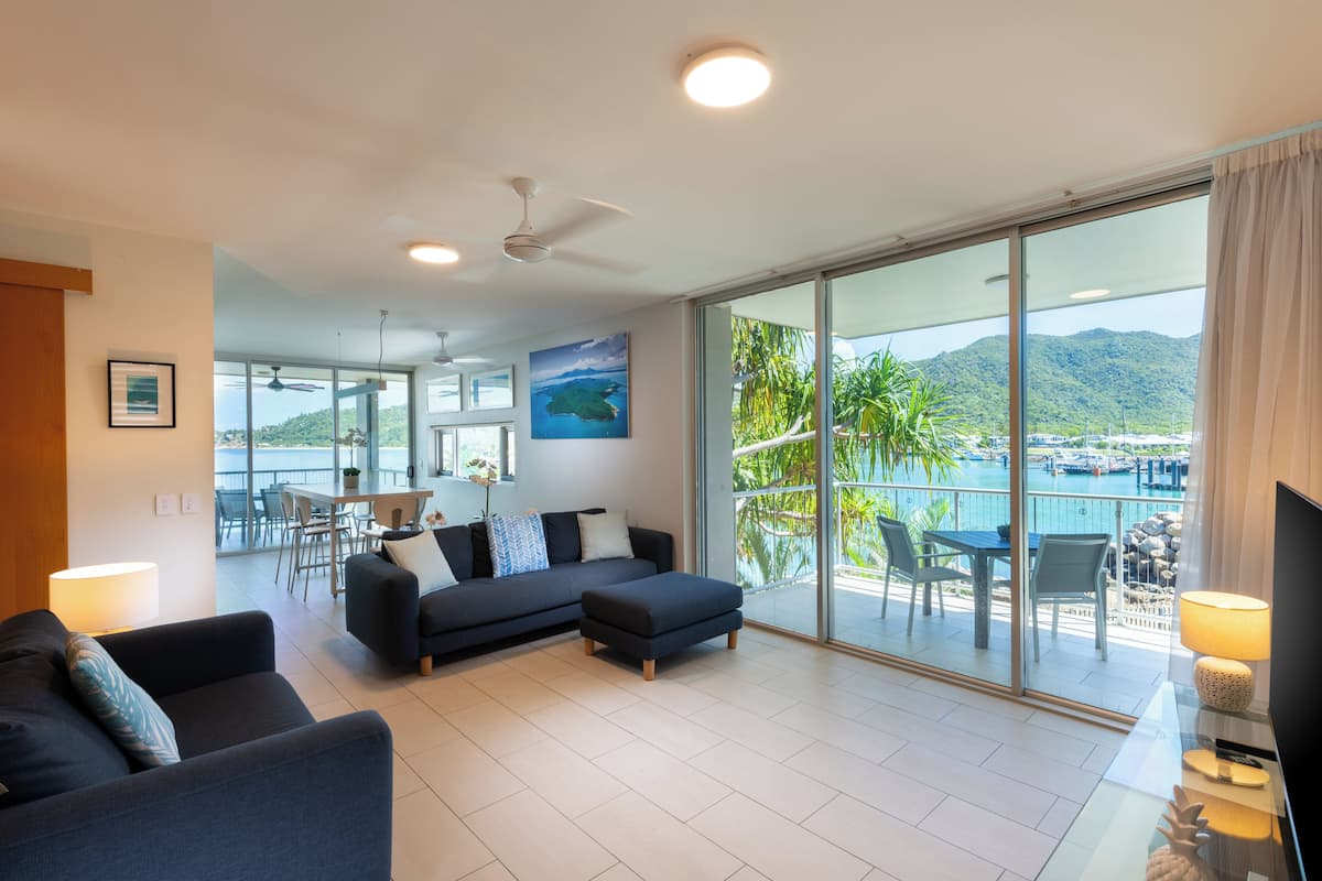 Ein modernes Wohnzimmer mit einer Couch, einem Hocker und einem Fernseher auf einem Glastisch. Große Fenster bieten Blick auf einen Pool und Berge.