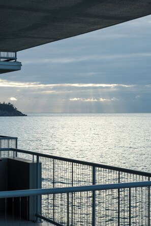 Une vue sur la mer depuis un balcon doté d’une rambarde en métal, avec une structure en béton au-dessus et une côte lointaine à l’horizon.