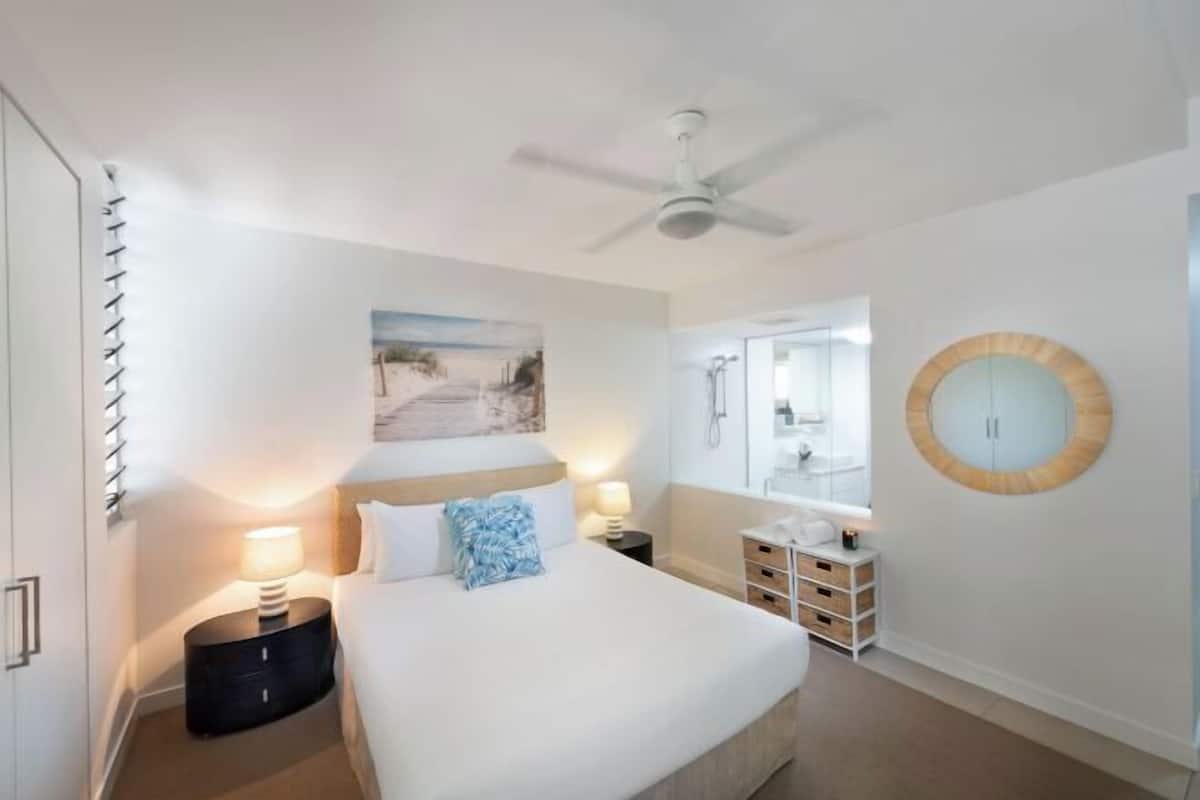 Ein Hotelzimmer mit Bett, Nachttischen, Deckenventilator, rundem Spiegel an der Wand und angrenzendem Badezimmer.