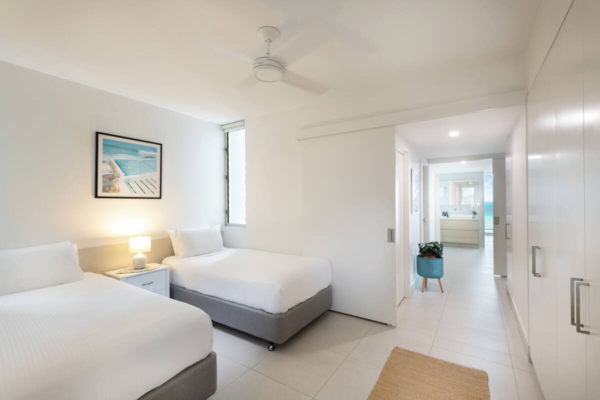 Ein modernes Hotelzimmer mit zwei Betten, einem Deckenventilator, einem Strandbild und Blick ins Badezimmer.