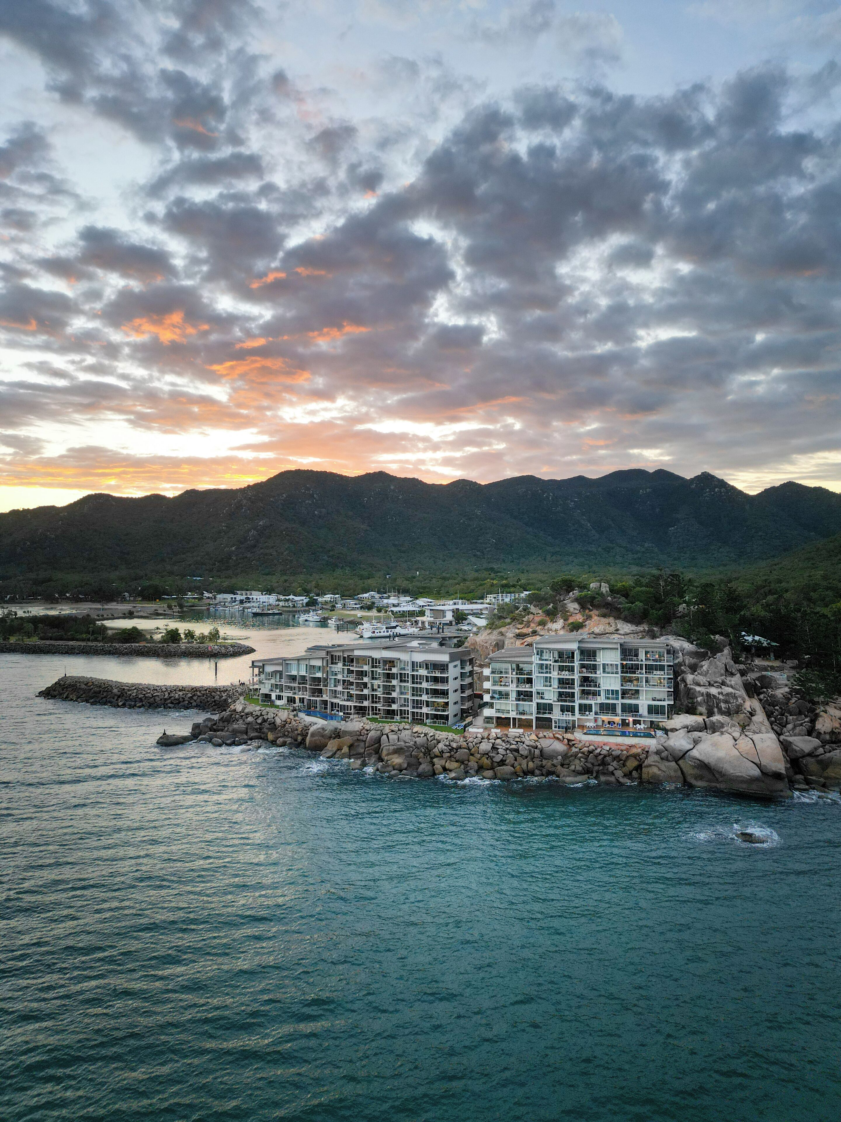 A coastal hotel with multiple buildings, a rocky shoreline, and mountains in the background.