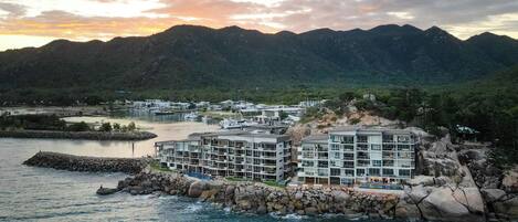 A coastal hotel with multiple buildings, a rocky shoreline, and mountains in the background.