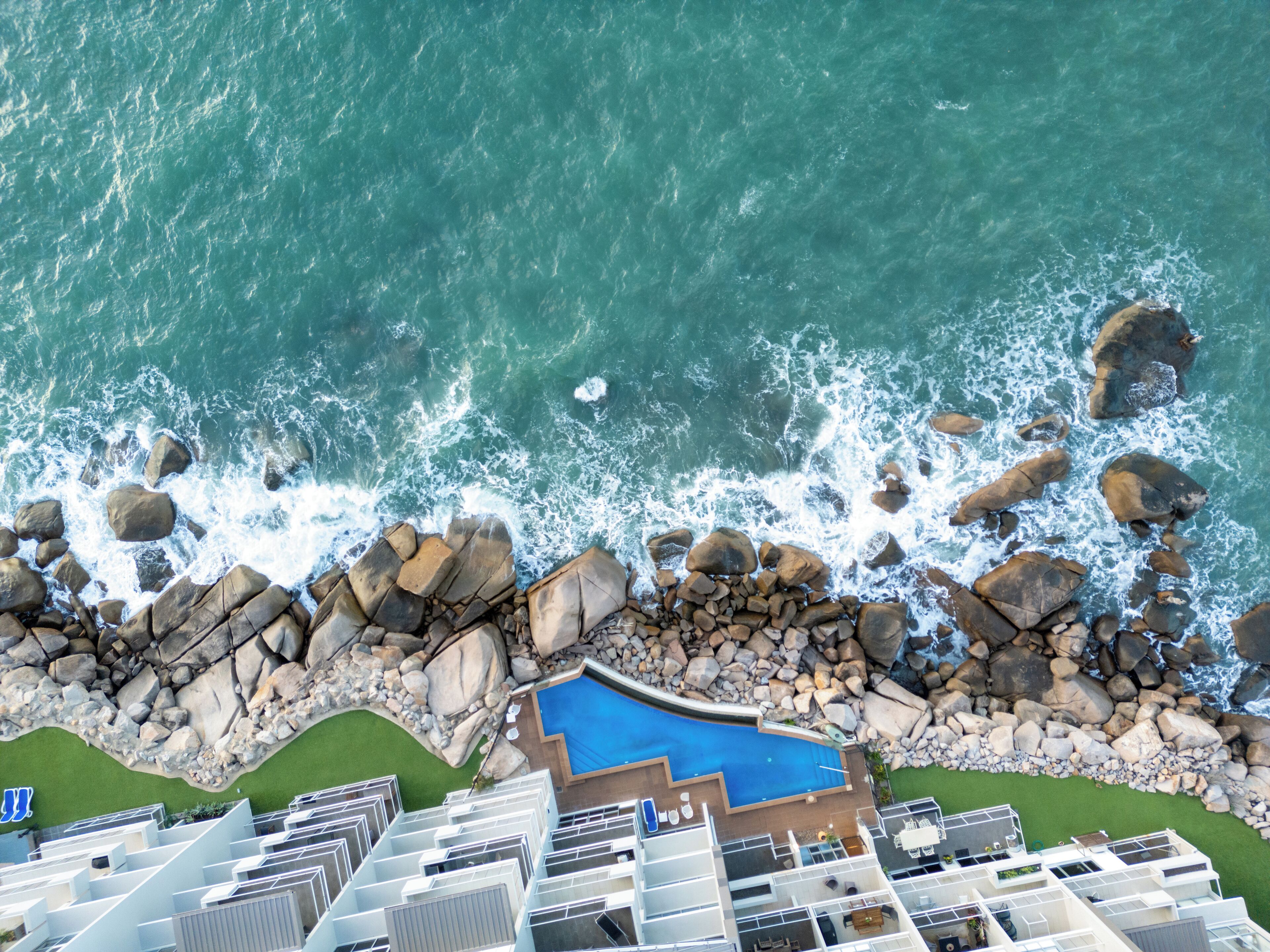 An aerial view of a coastal hotel with a swimming pool surrounded by rocks and waves.