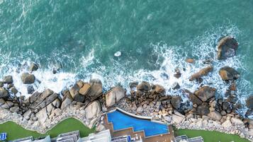 An aerial view of a coastal hotel with a swimming pool surrounded by rocks and waves.
