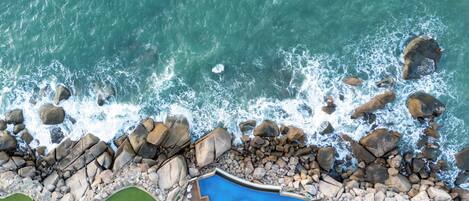 Vista aérea de un hotel costero con piscina, rodeado de rocas y olas.