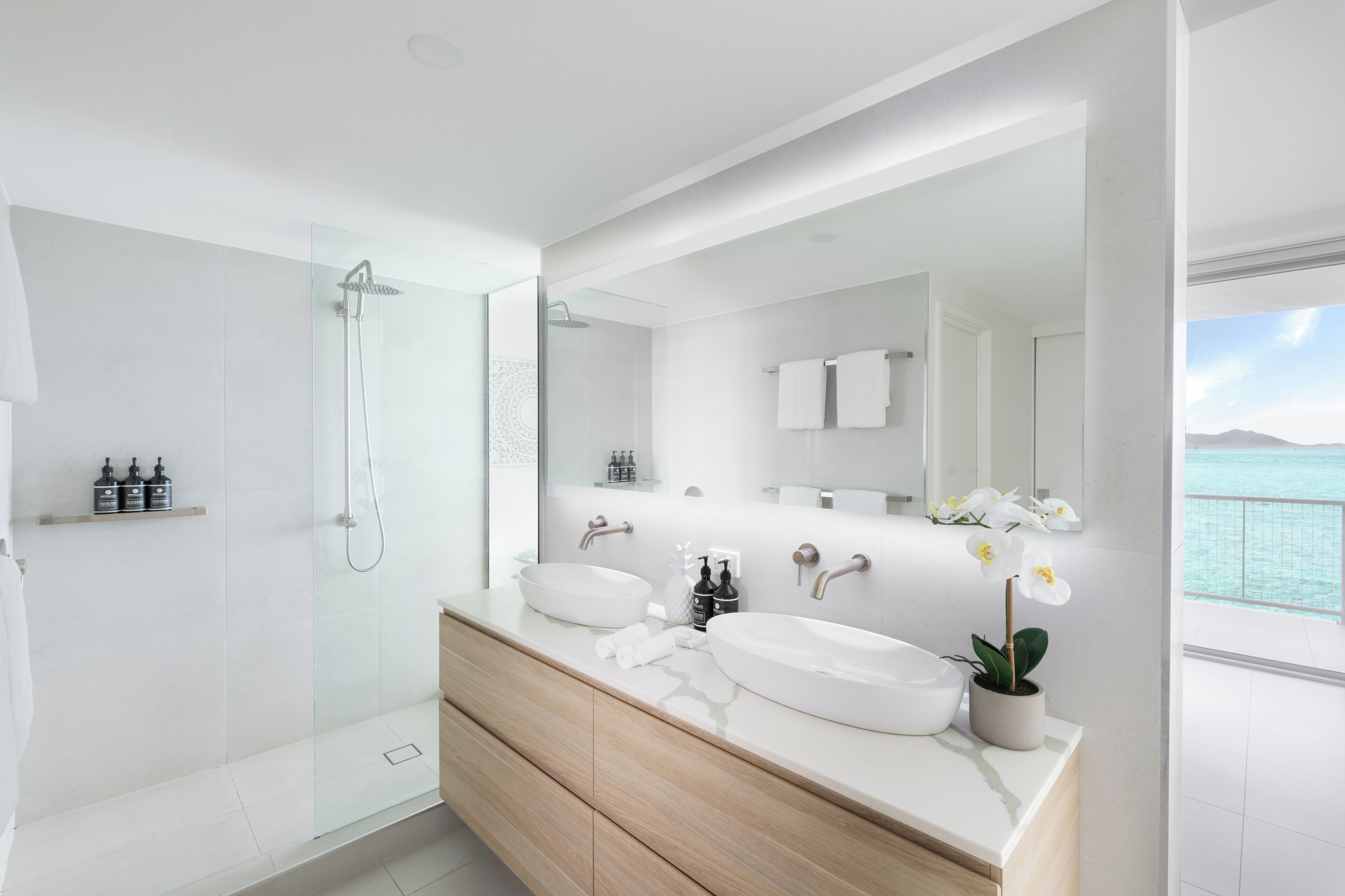 A modern bathroom with a glass-enclosed shower, a large mirror, and a view of the sea.