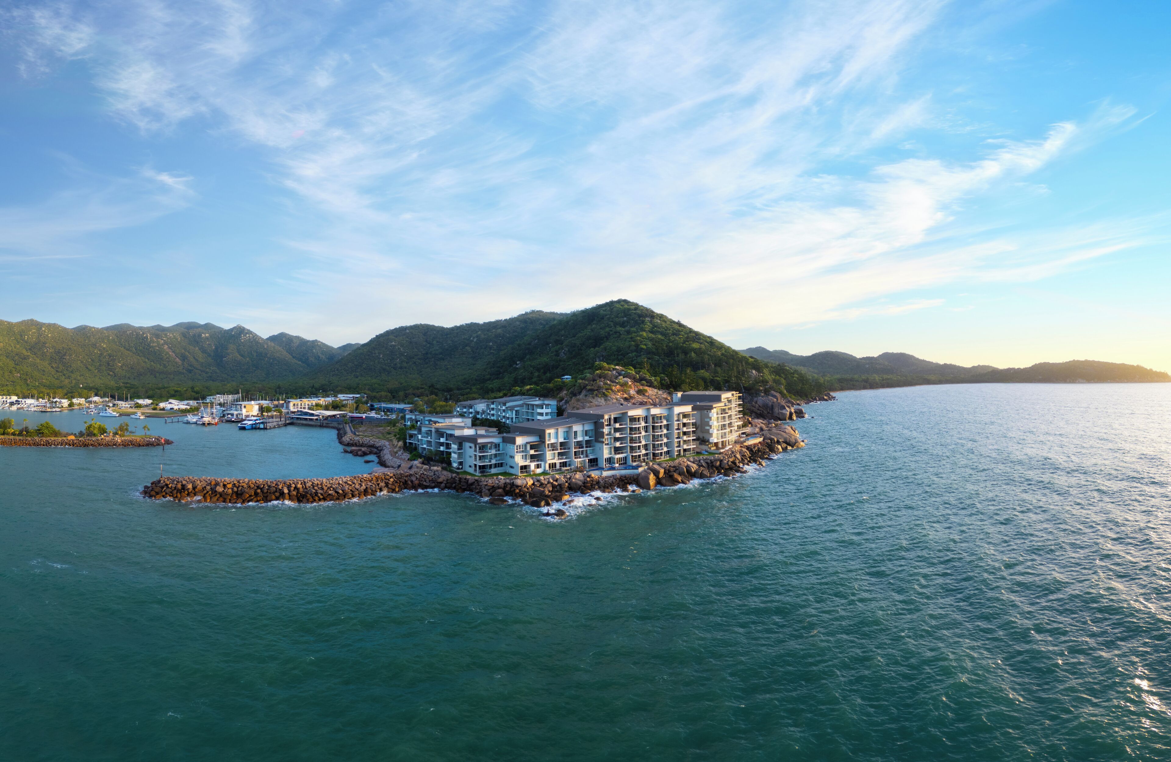 A coastal hotel with multiple buildings, a rocky breakwater, and a mountainous backdrop.