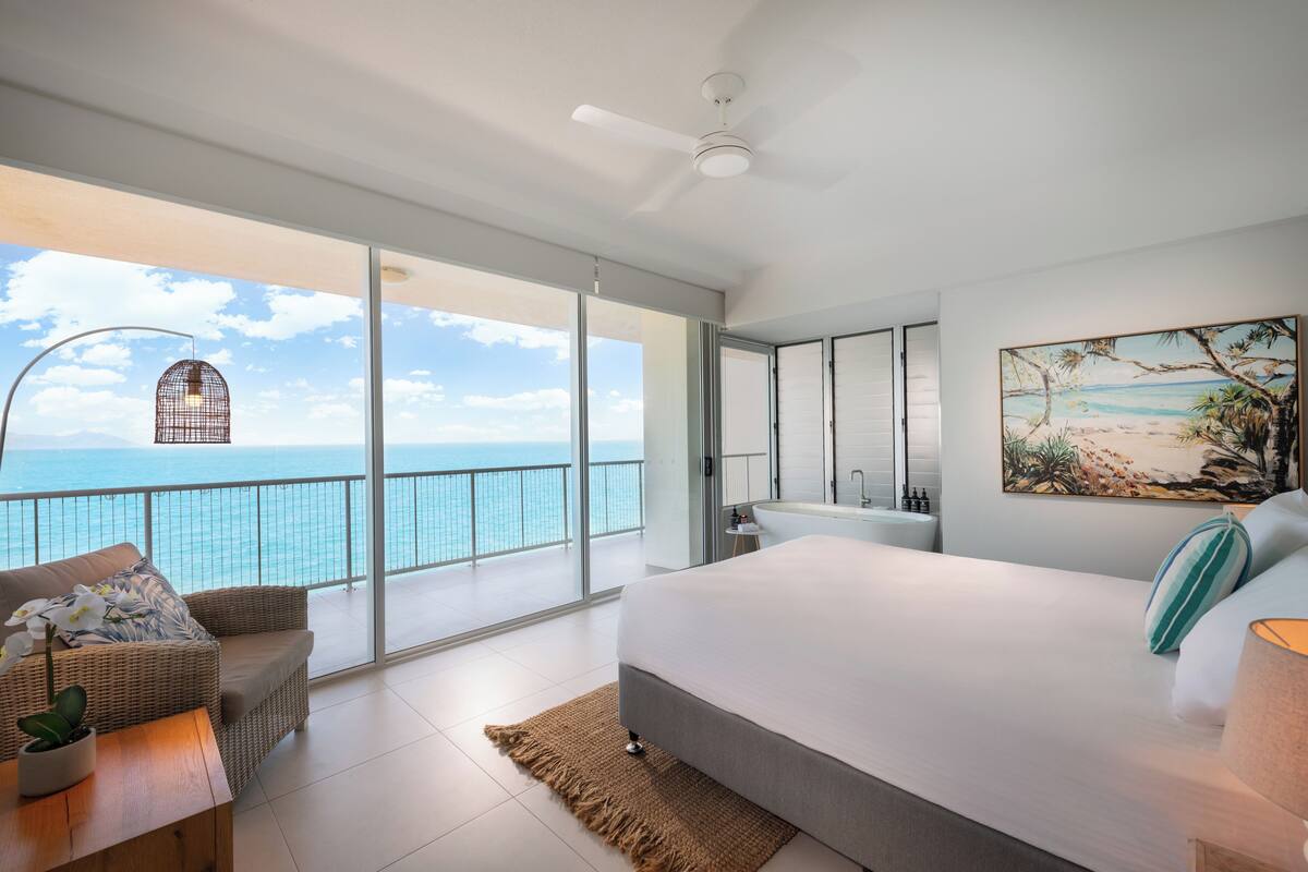 Ein geräumiges Schlafzimmer mit einem großen Bett, einem Balkon mit Meerblick sowie ein Badezimmer mit großem Spiegel und Waschbecken.