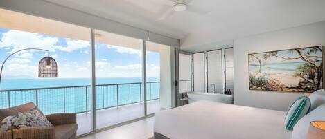 A spacious bedroom with a large bed, a balcony with a view of the ocean, and a bathroom with a large mirror and sink.