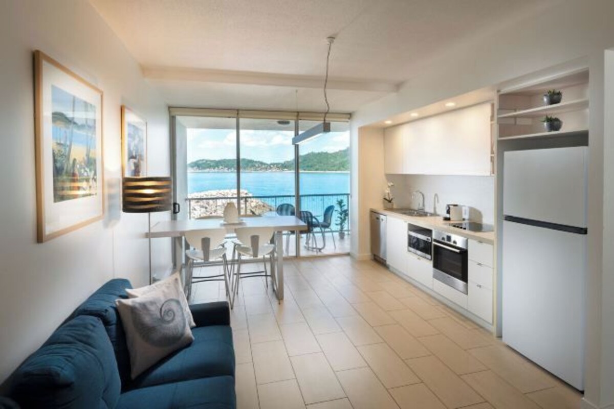 A modern kitchen with white cabinets and stainless steel appliances, a dining area with a table and chairs, and a sofa with a cushion.