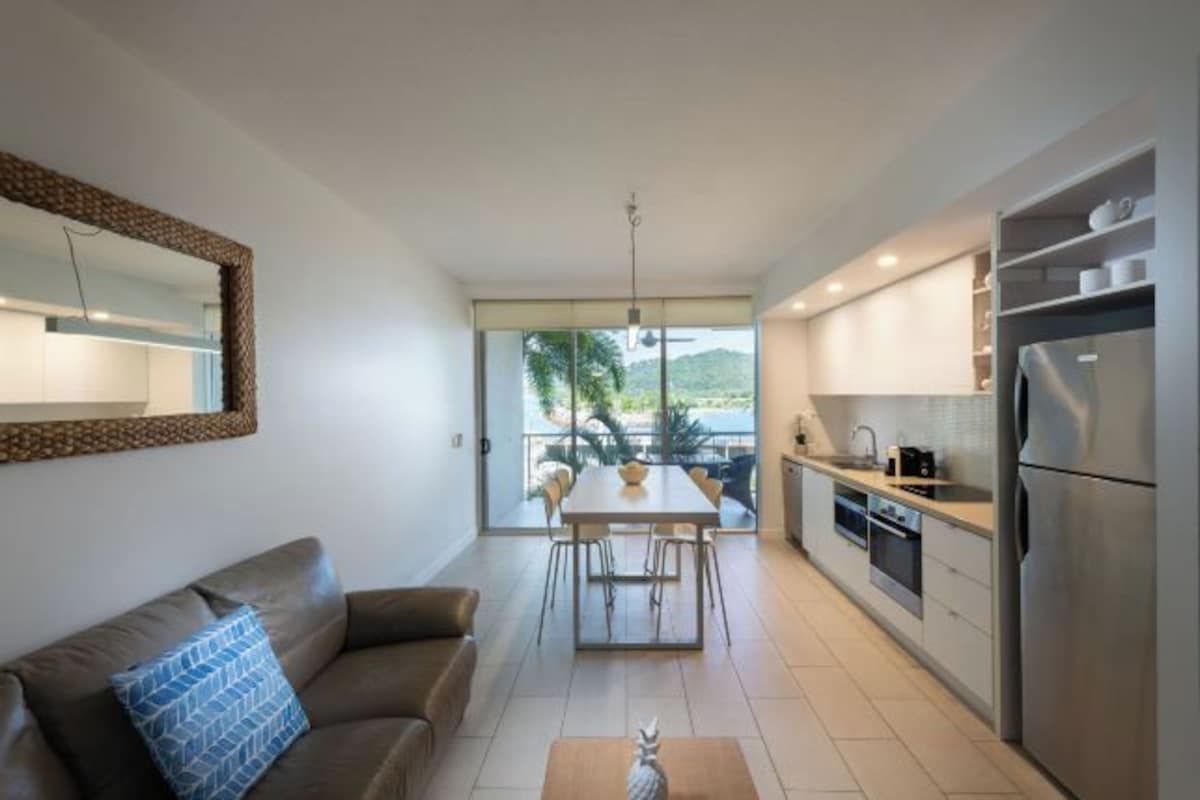 A modern kitchen with a dining area, a sofa, and a large window offering a view of greenery.