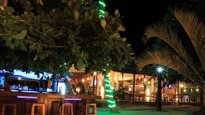 Uma área de bar com bancos de madeira, balcão e uma palmeira com luzes verdes.