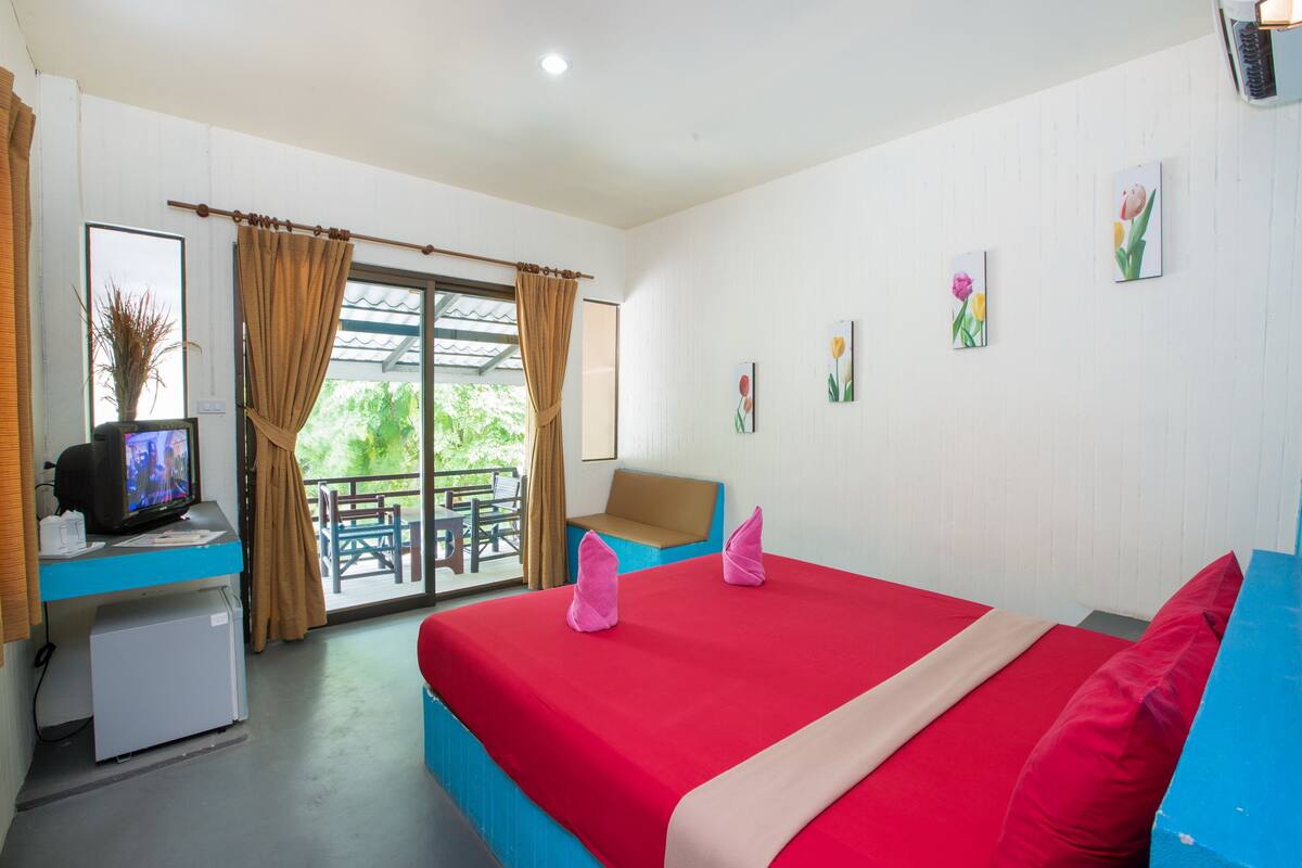 Quarto de hotel com cama, escrivaninha com televisão, mini-geladeira e varanda com cadeira.