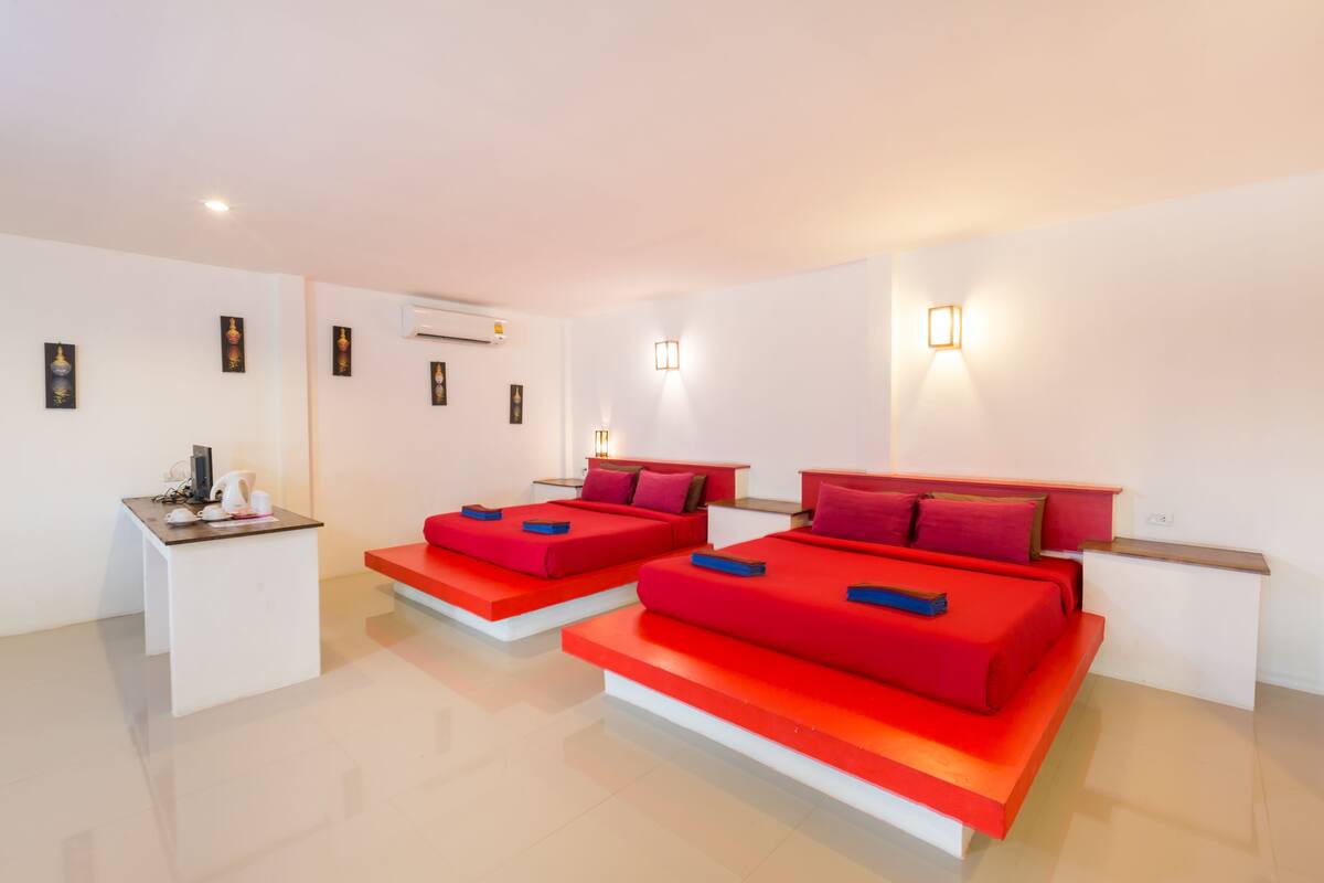 Quarto de hotel moderno com duas camas vermelhas, uma pequena escrivaninha e um ar condicionado branco na parede.