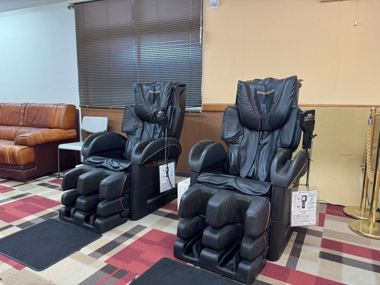 Two massage chairs in a room with a checkered carpet, a brown leather sofa, and a window with blinds.
