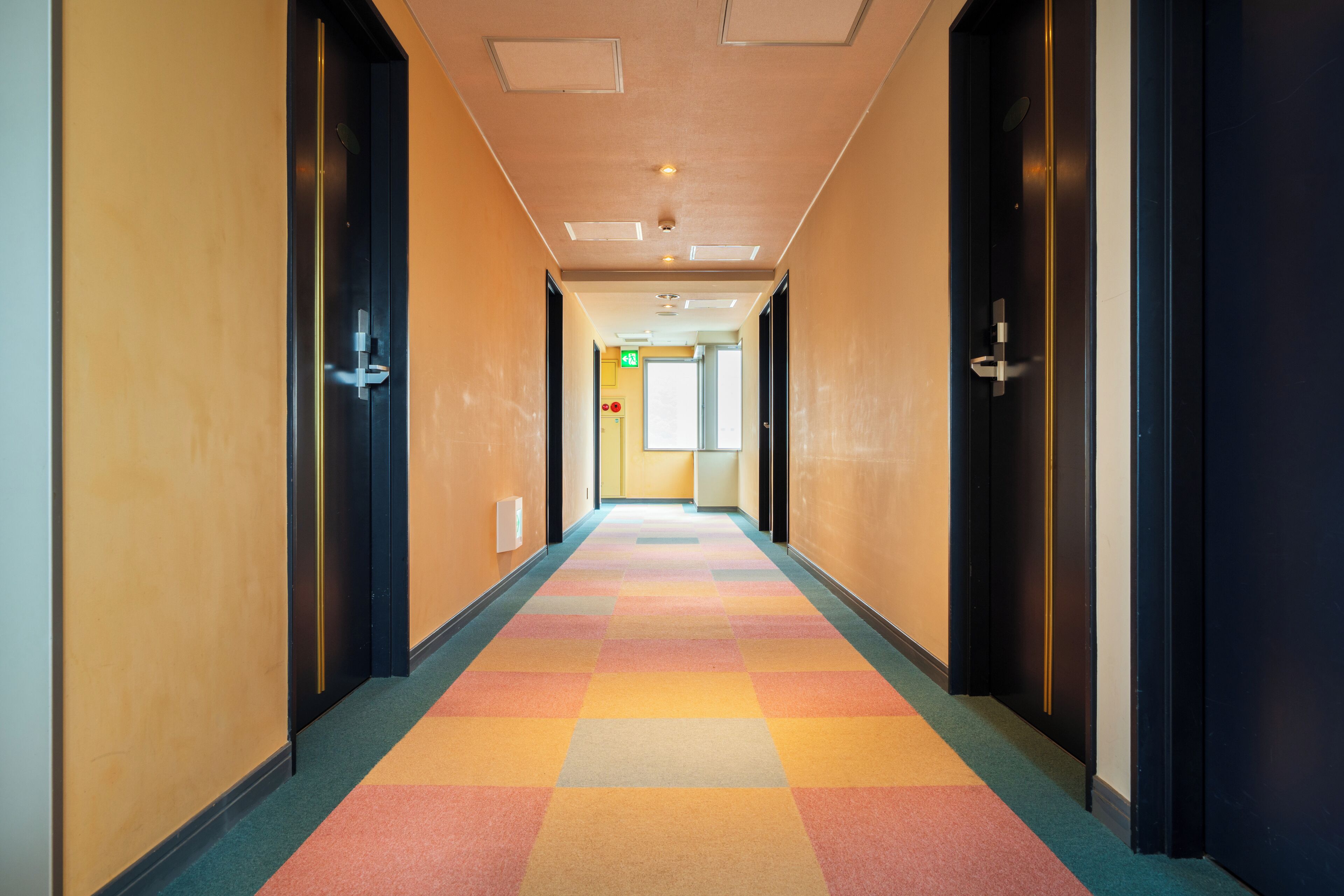 Pasillo de hotel con una alfombra colorida, puertas a ambos lados y una señal de salida visible.