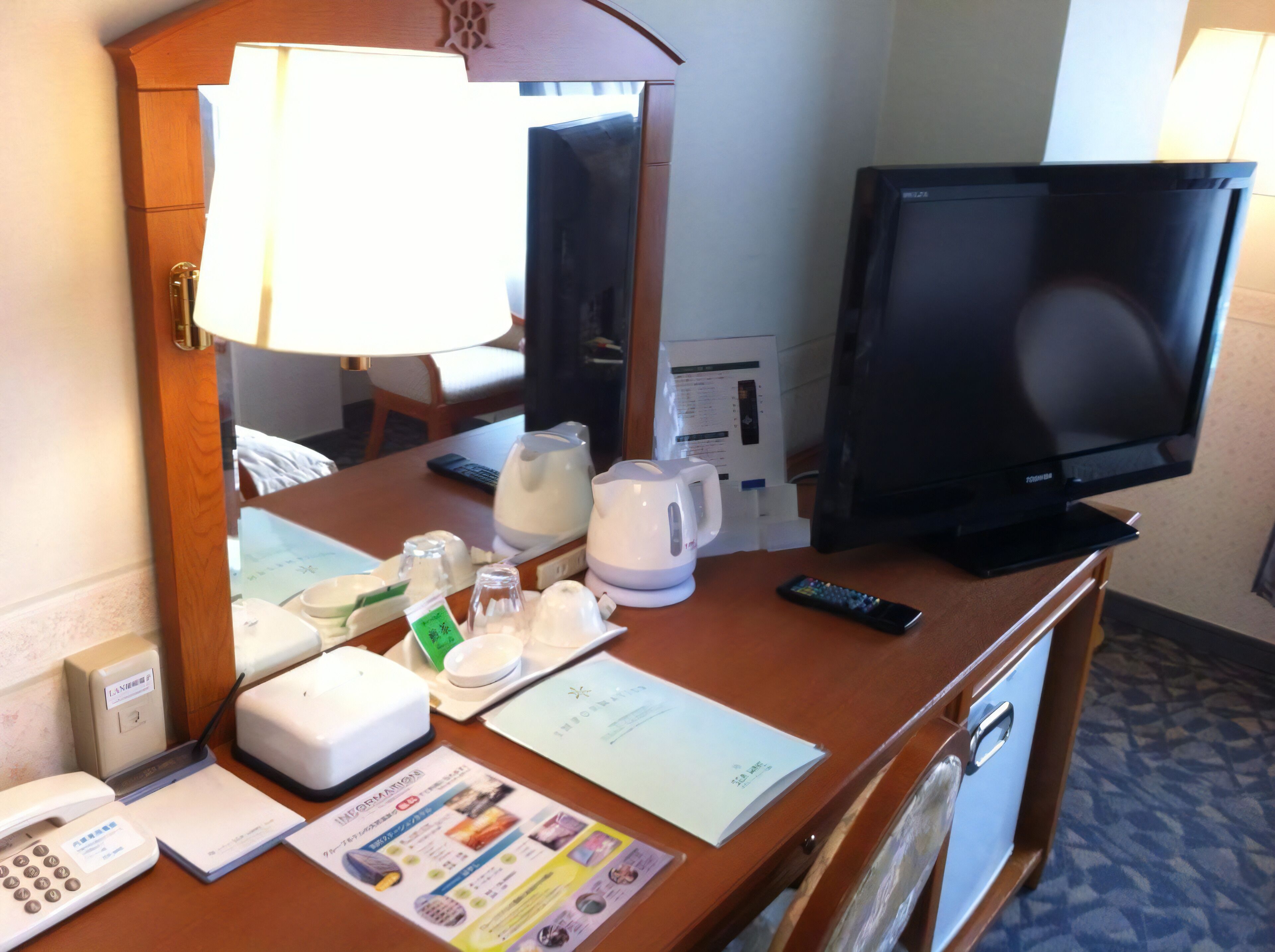Ein Schreibtisch im Hotelzimmer mit Fernseher, Lampe, Telefon, Wasserkocher und einigen Informationsbroschüren.