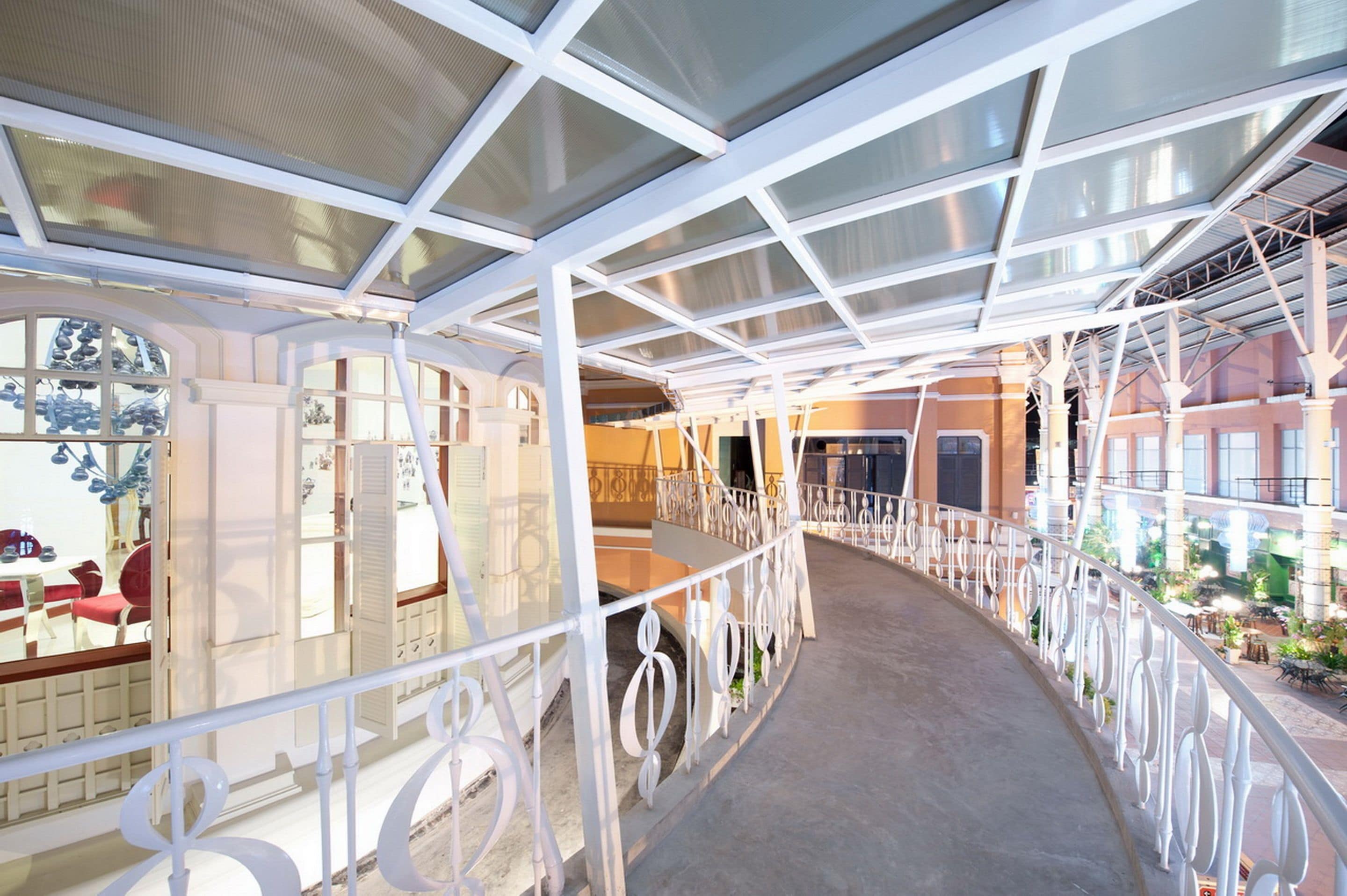 A covered walkway with white railings and glass panels, leading to a building with a red facade and outdoor seating.