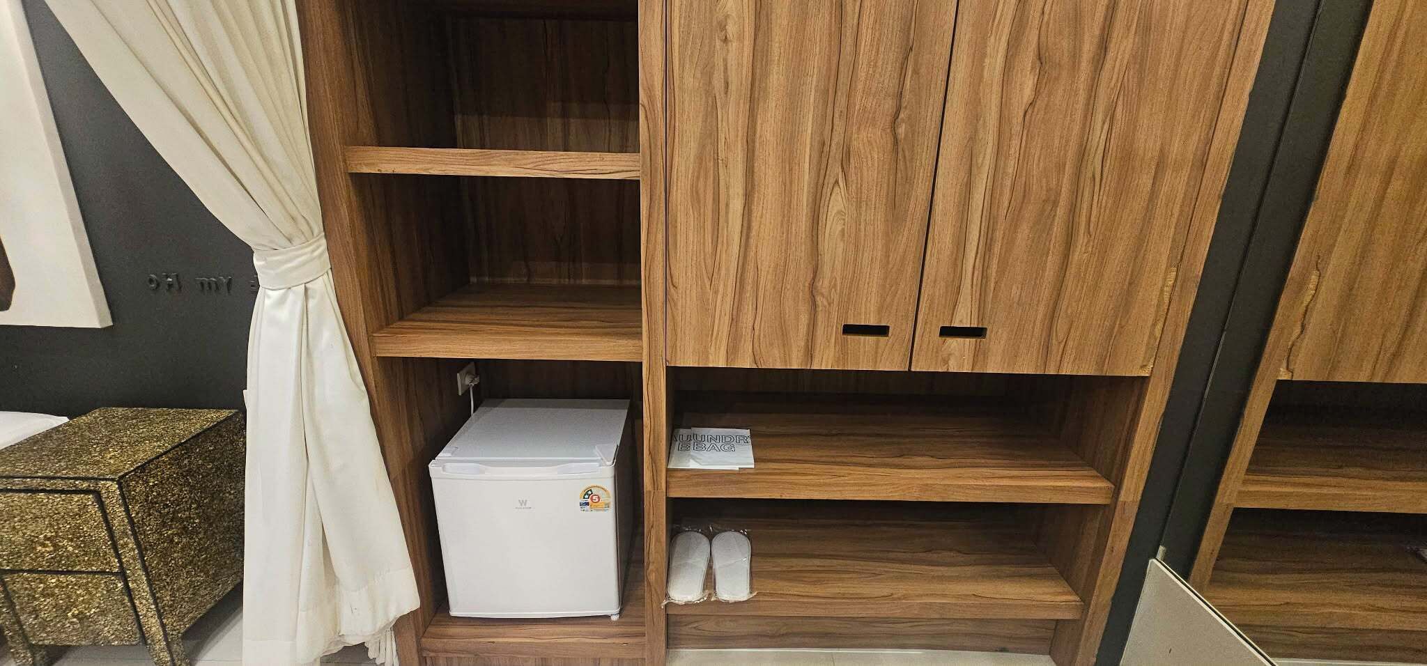 A wooden wardrobe with multiple shelves and a small white appliance, likely a mini-fridge, in a room with a dark wall and a curtain.