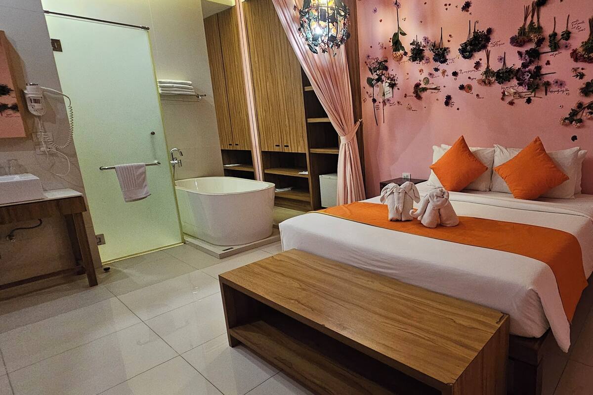 A bedroom with a bed, two pillows, a wooden bench, a bathroom with a bathtub, and a shower curtain.