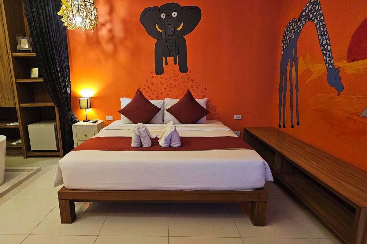 A bedroom with a bed, pillows, a nightstand, a lamp, a wall mural, a bathtub, and a wooden bench.