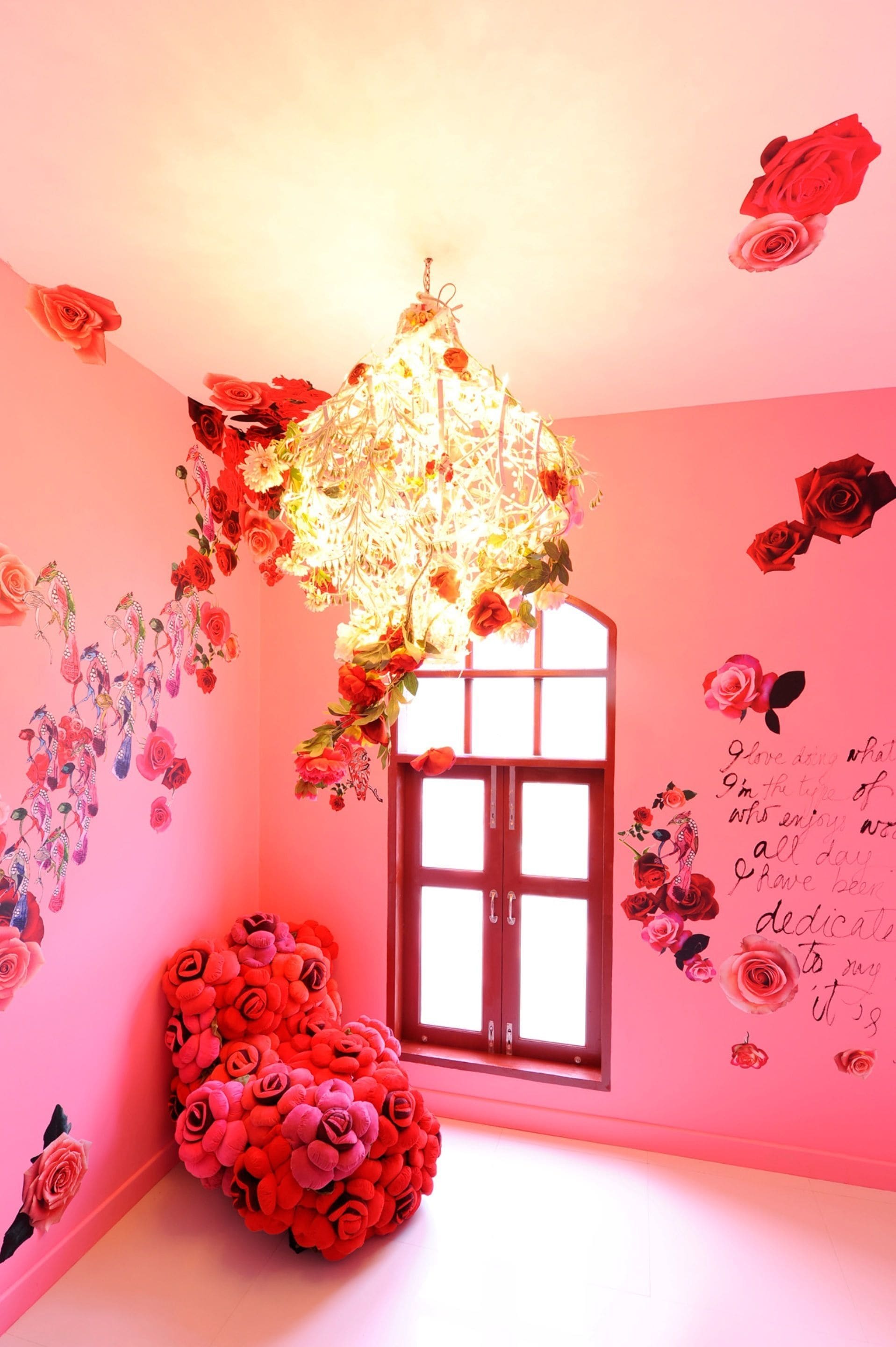 A room with pink walls, a large window, and various floral decorations including a chandelier, a heart-shaped arrangement of roses, and wall decals.