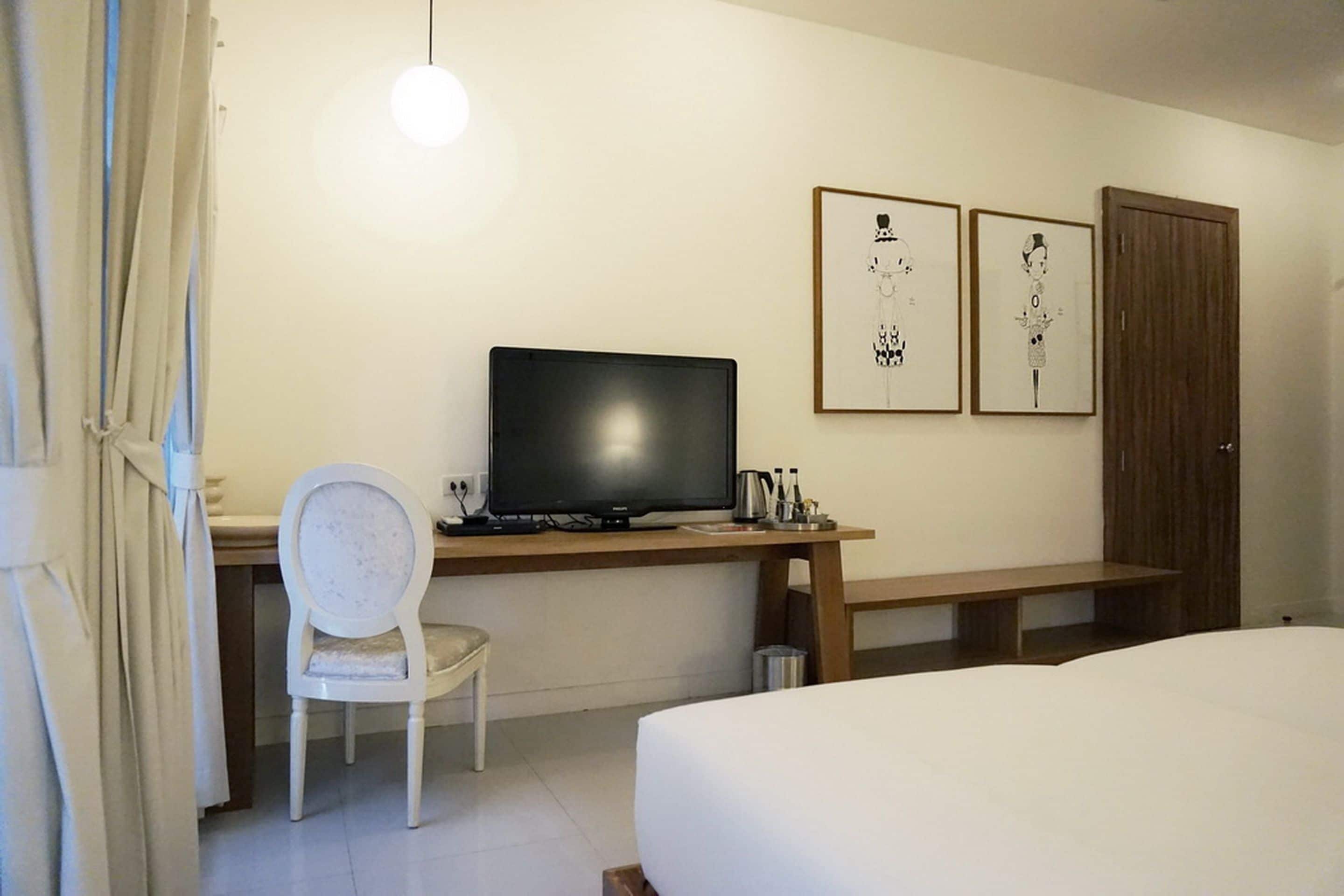 A hotel room with a bed, a desk with a computer, a chair, and two framed pictures on the wall.