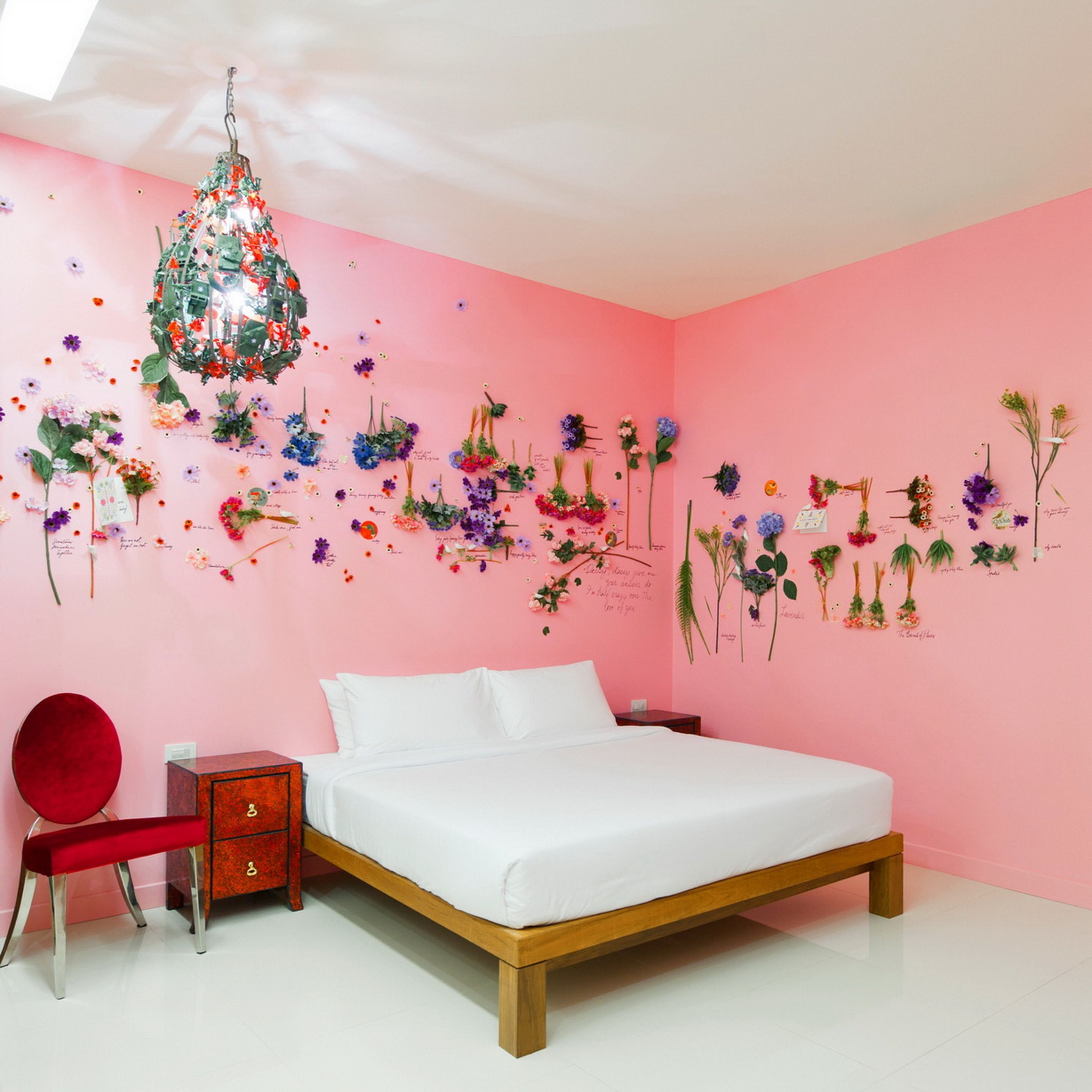 Un dormitorio con pared rosa, una cama de madera, una silla roja, una mesita de noche de madera y una araña.