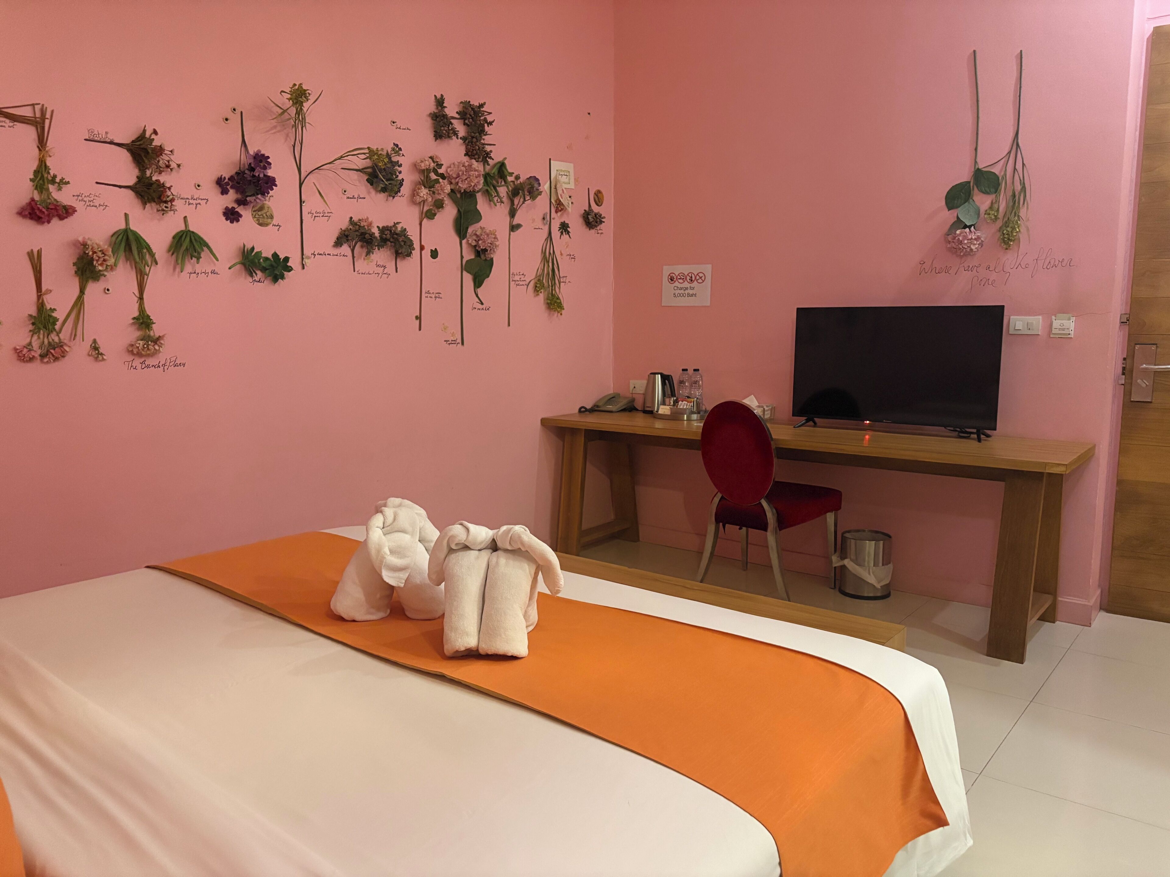 A hotel room with a bed, a desk with a TV, a chair, and a wall decorated with various plants.