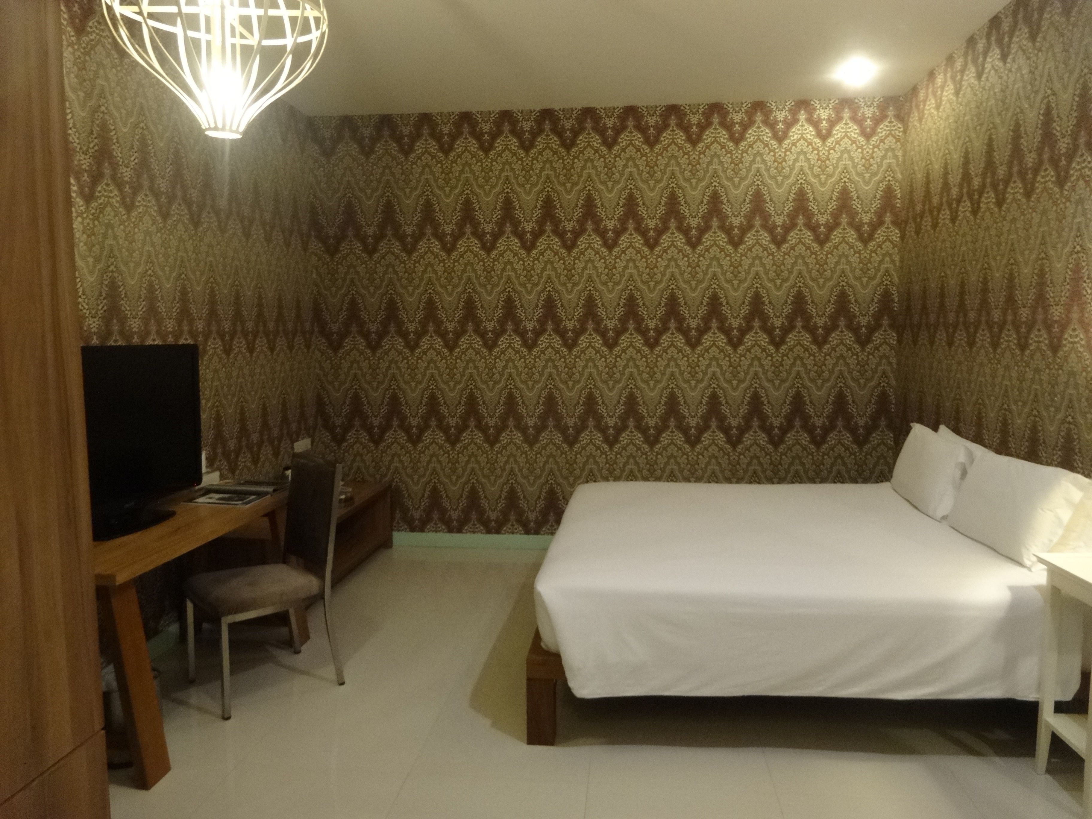 A hotel room with a bed, a desk with a chair, a television, and patterned wallpaper.