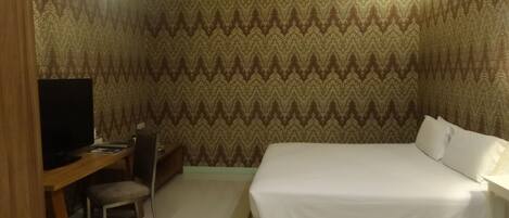 A hotel room with a bed, a desk with a chair, a television, and patterned wallpaper.