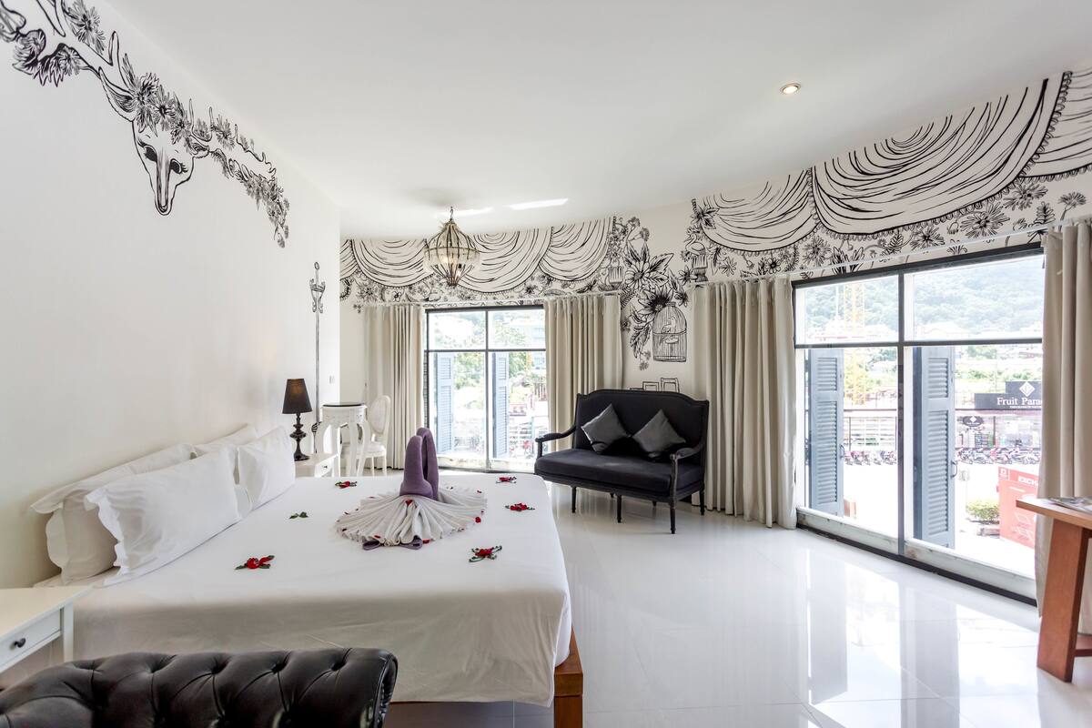 A spacious hotel room with a large bed, a black leather chair, and a view of the city through large windows.