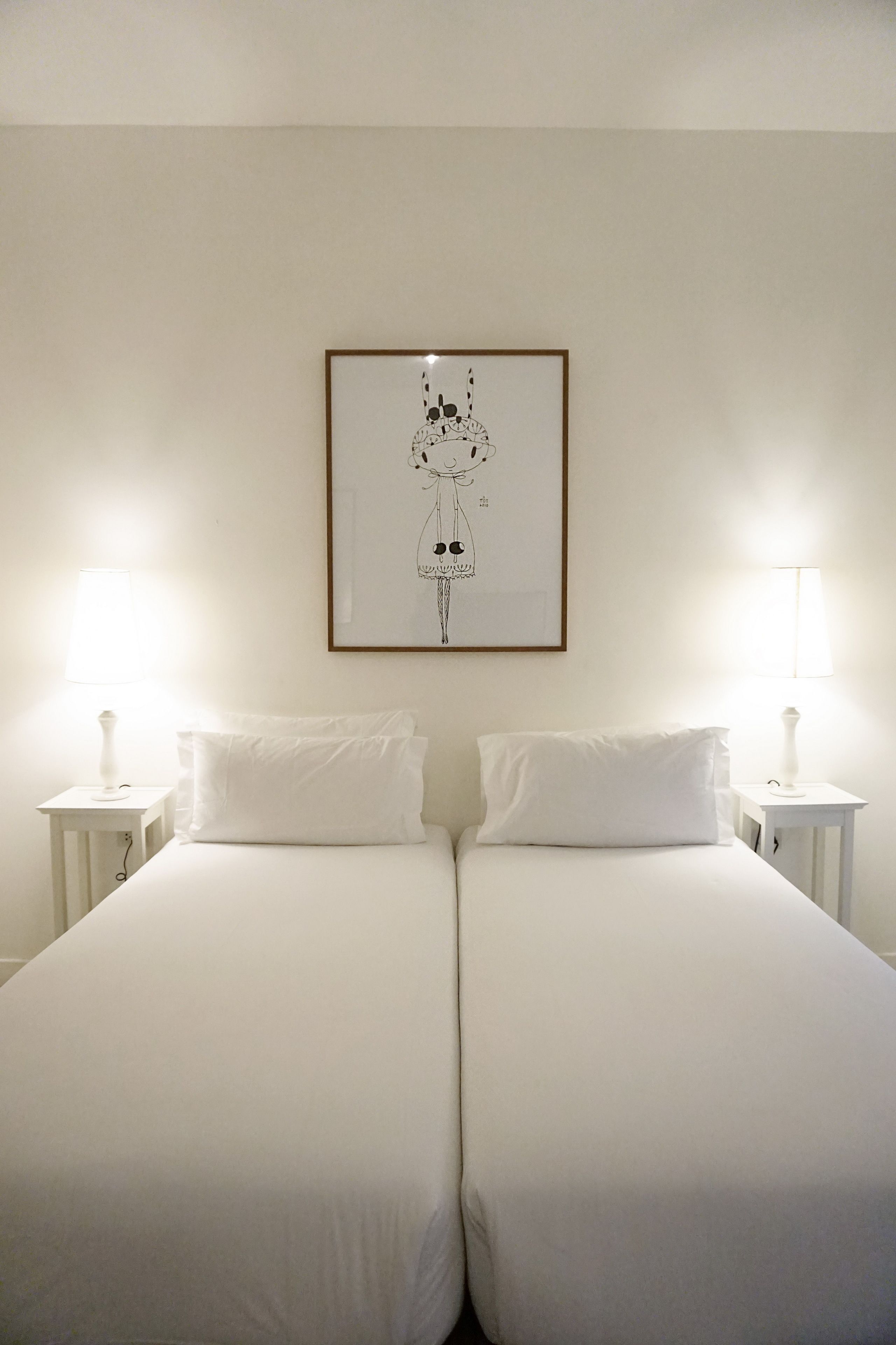A double bed with white linens, two bedside lamps, and a framed picture on the wall.