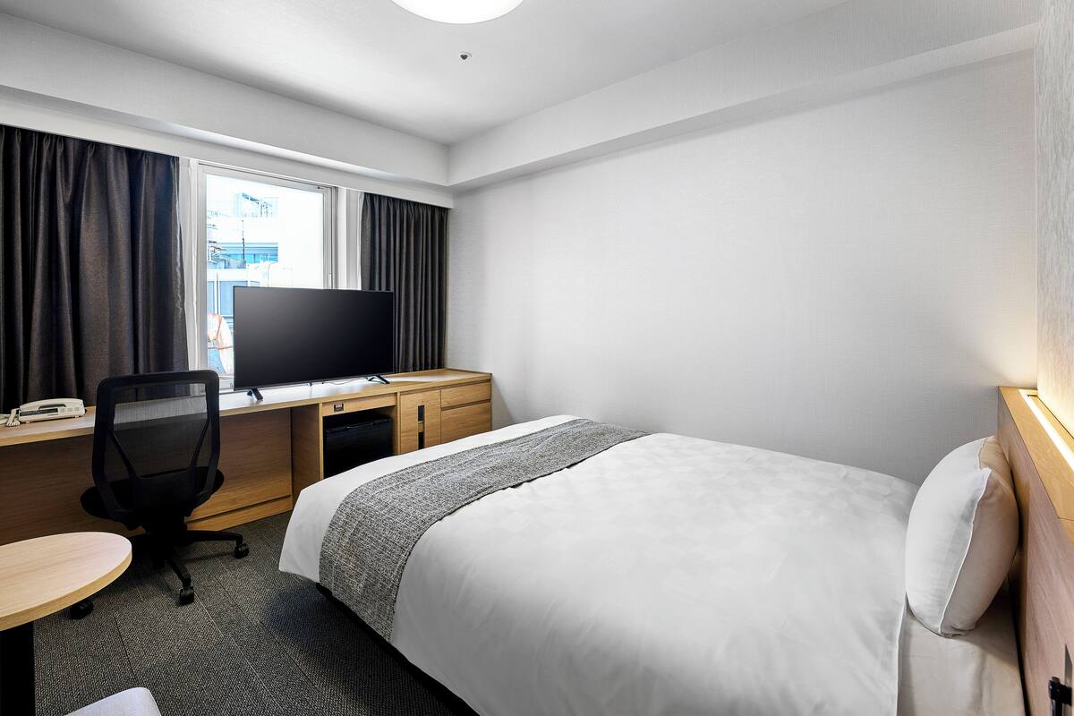 Habitación de hotel con cama, escritorio, computadora, silla y ventana con cortinas.