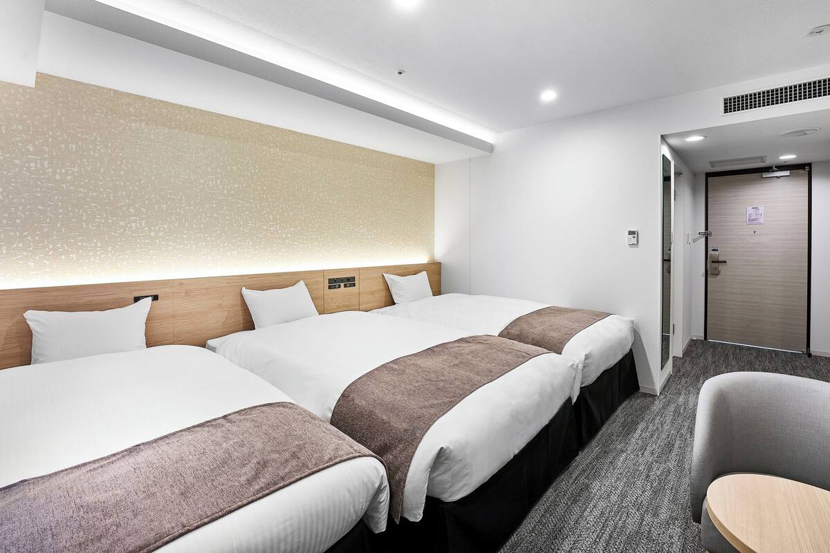 Habitación de hotel con tres camas, cabecera de madera, luz montada en la pared y una puerta con un letrero.