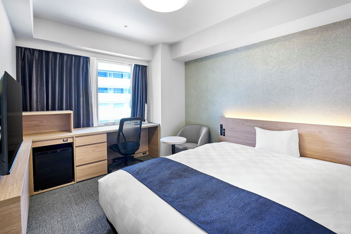 Una habitación de hotel con cama, escritorio, silla, televisor y ventana con cortinas.