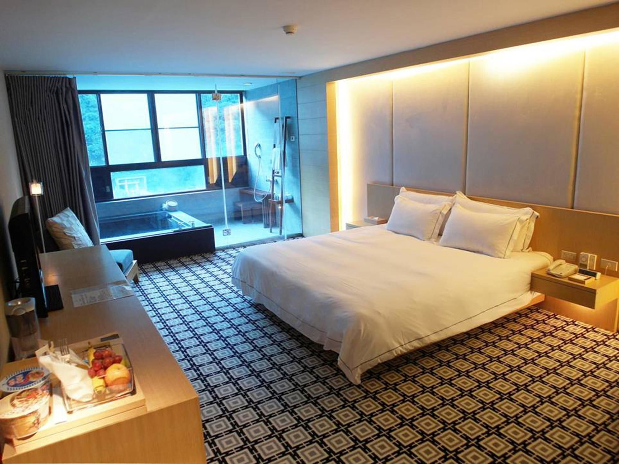 A hotel room with a large bed, a desk with a chair, a small table with fruits, and a patterned floor.