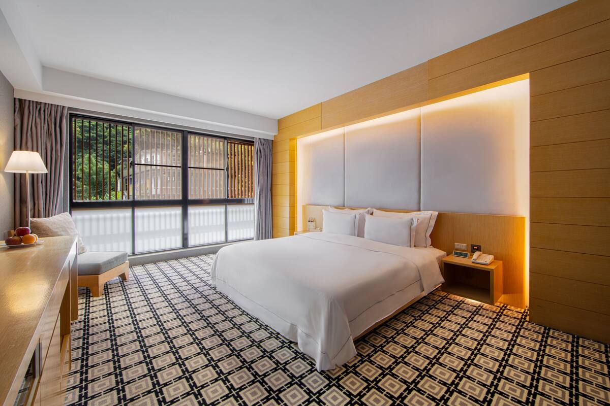 A hotel room with a large bed, a desk with a chair, a nightstand with a phone, and a window with curtains.